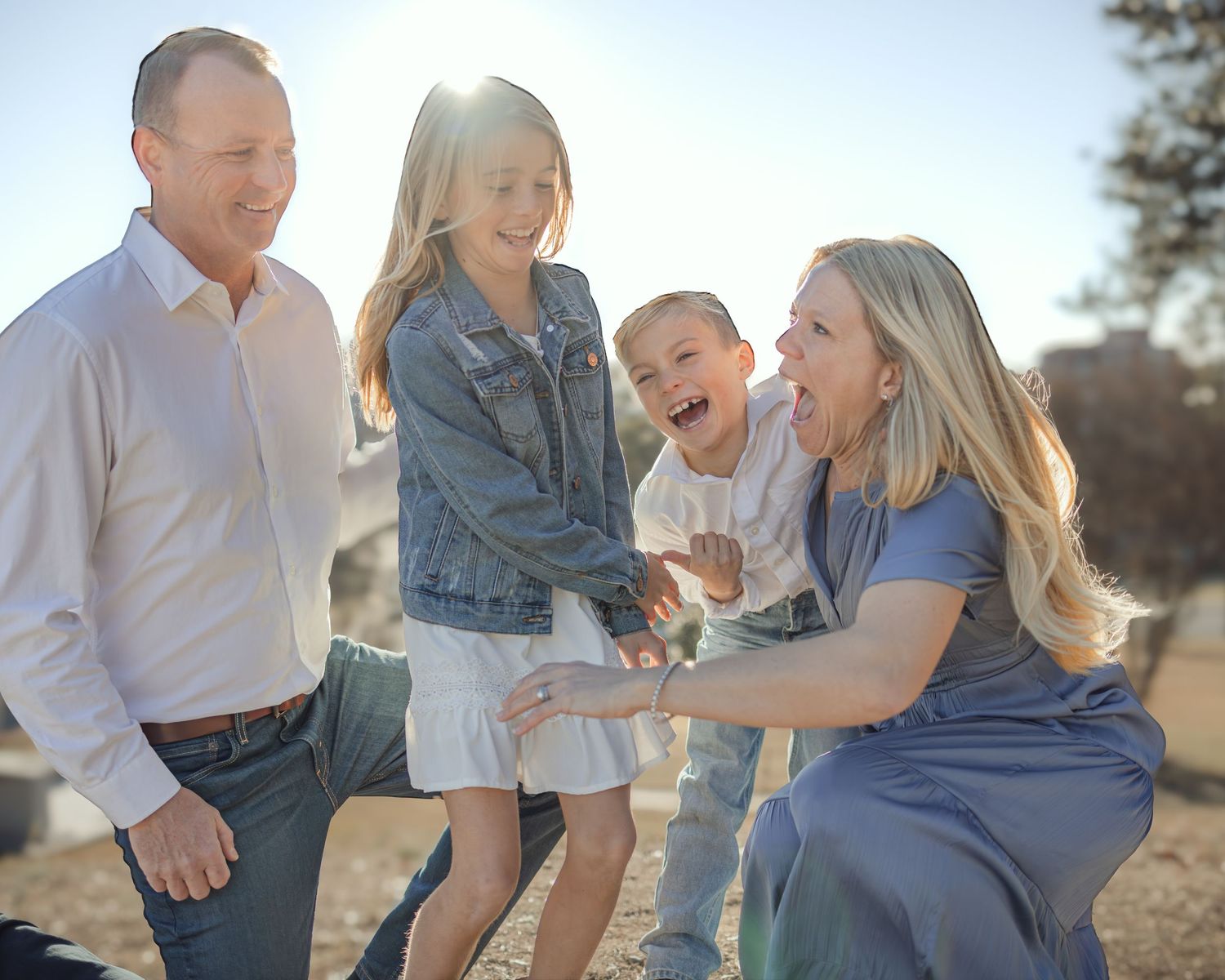 Happy family of four laughing together outdoors in golden sunlight, parents and two young children sharing a joyful moment.