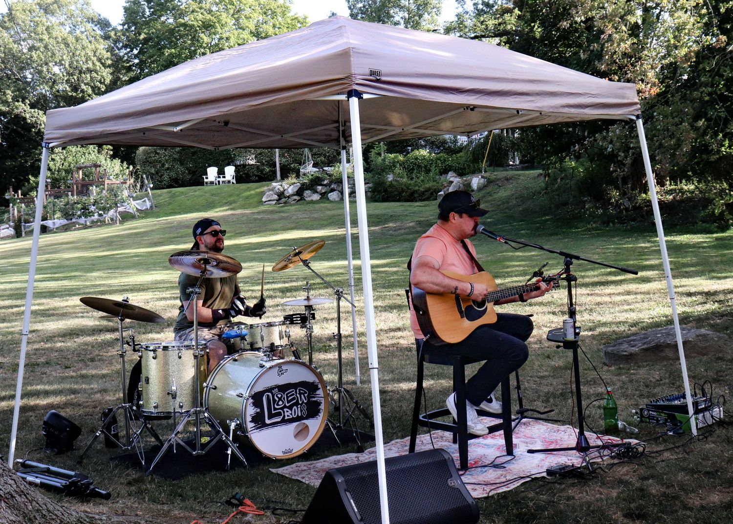 Musicians perform with guitars and drums under white canopy tents in a shaded outdoor park setting.