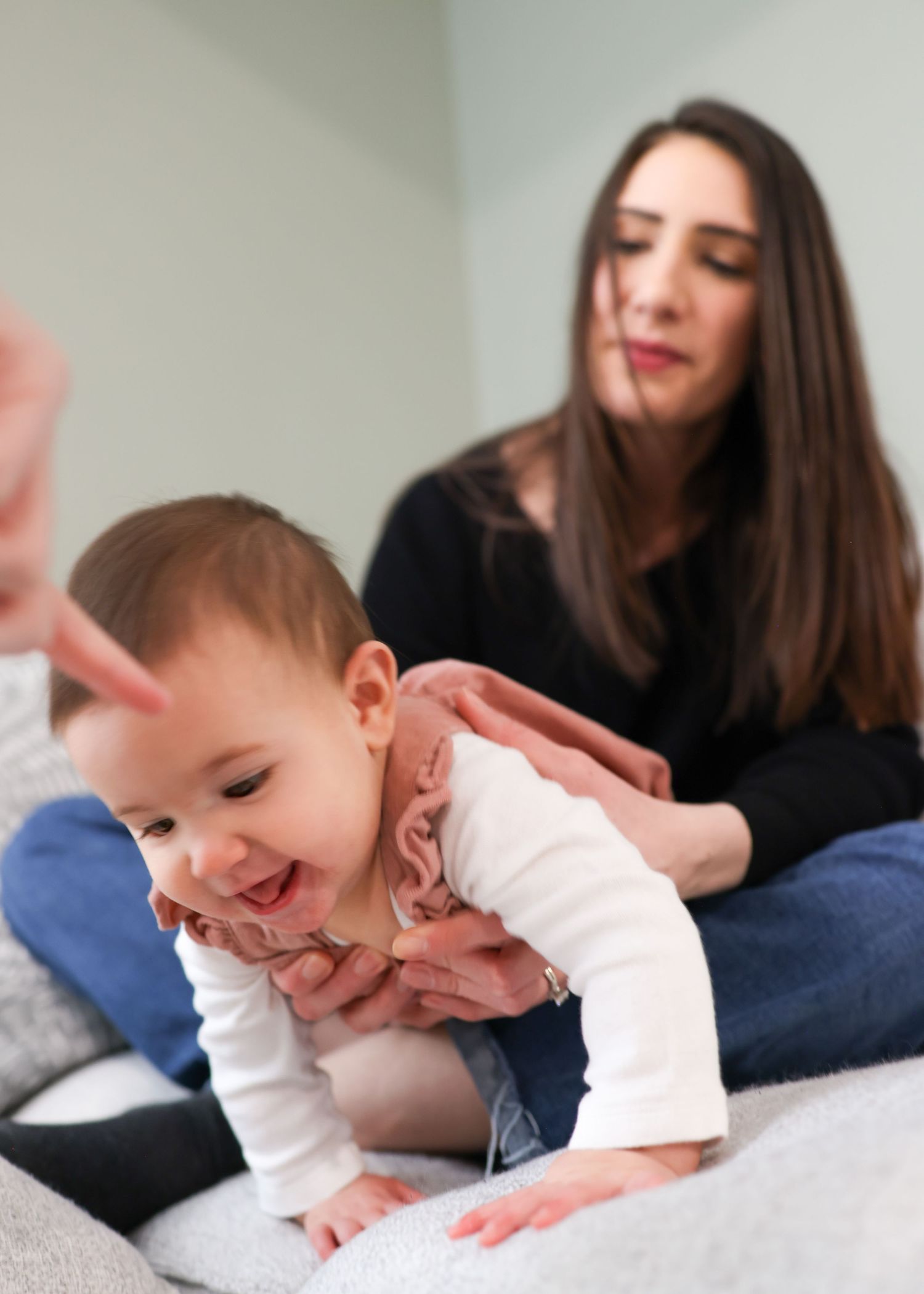 A smiling baby crawls on a bed while a dark-haired woman in black sits behind, gently supporting the child.