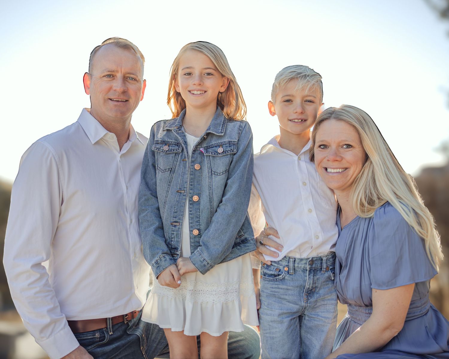 A family wearing coordinated blue and white outfits poses together for outdoor portrait photos at sunset.
