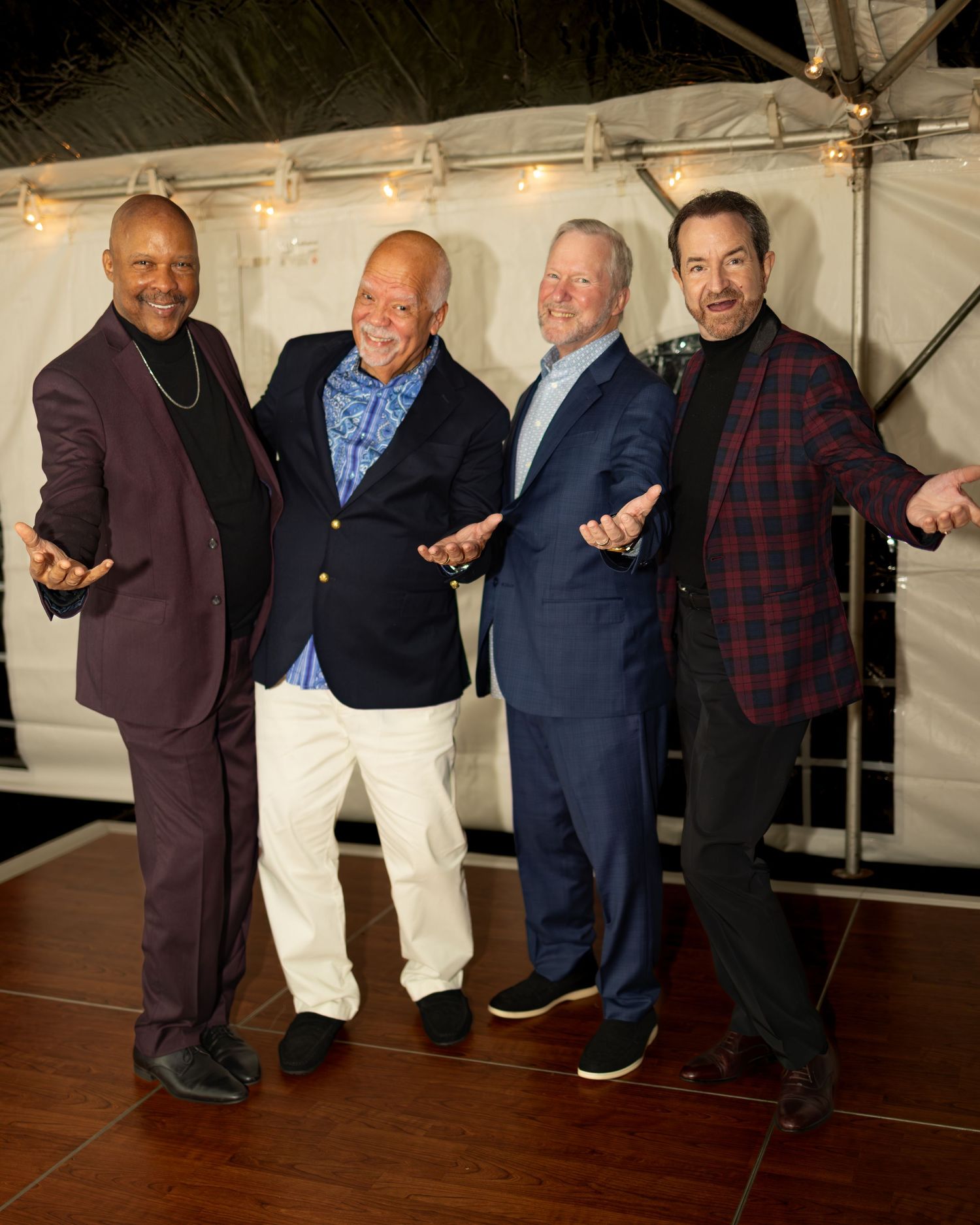 Four smiling men pose together at an indoor event, wearing stylish blazers and casual attire under string lights.