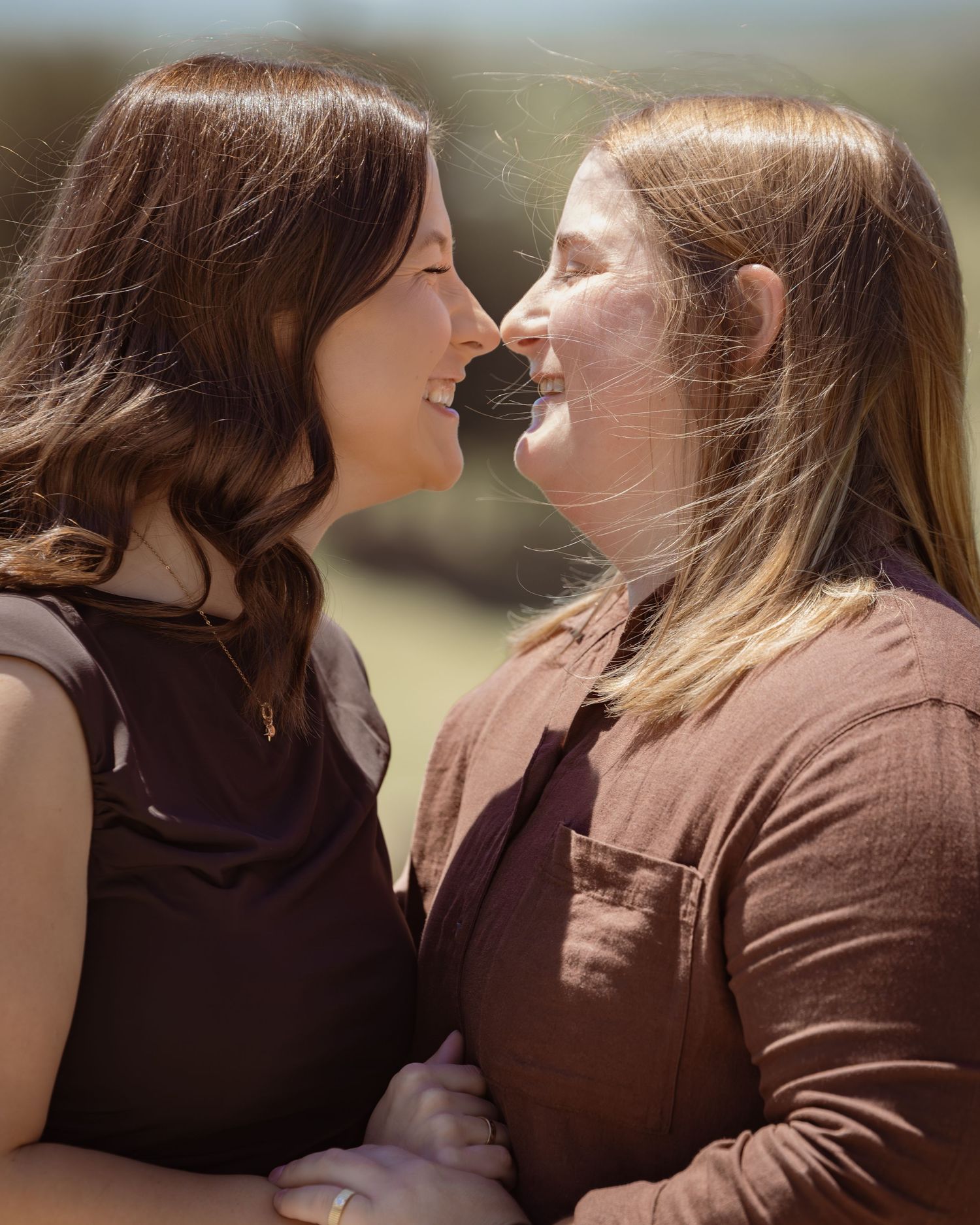 Two women smiling and sharing an intimate moment outdoors, nearly touching noses in a romantic pose.