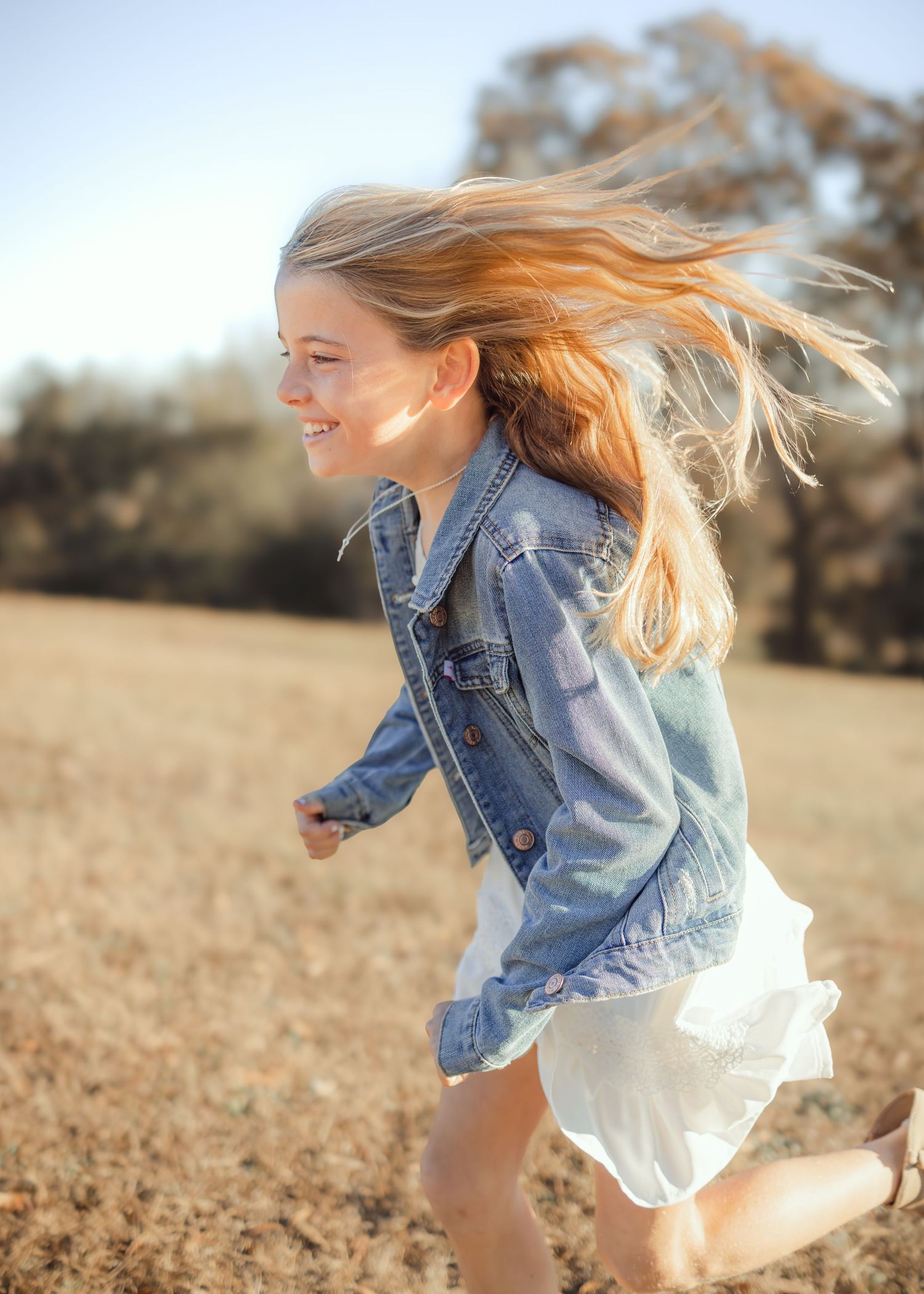 A joyful young girl with flowing blonde hair runs through a golden field wearing a white dress and denim jacket.