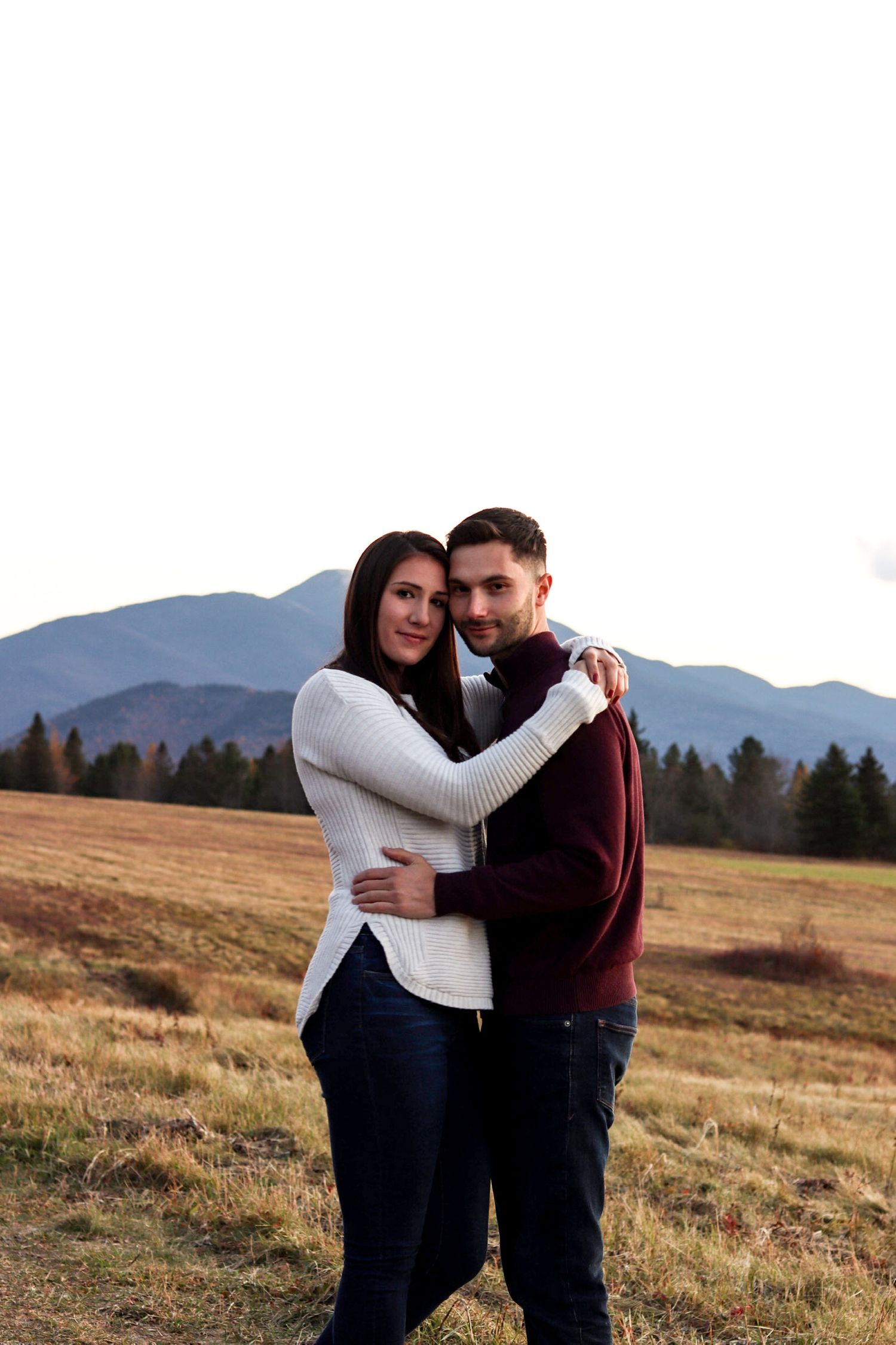 A couple embracing in an open field with mountain and forest backdrop, wearing cozy sweaters in a romantic outdoor portrait.