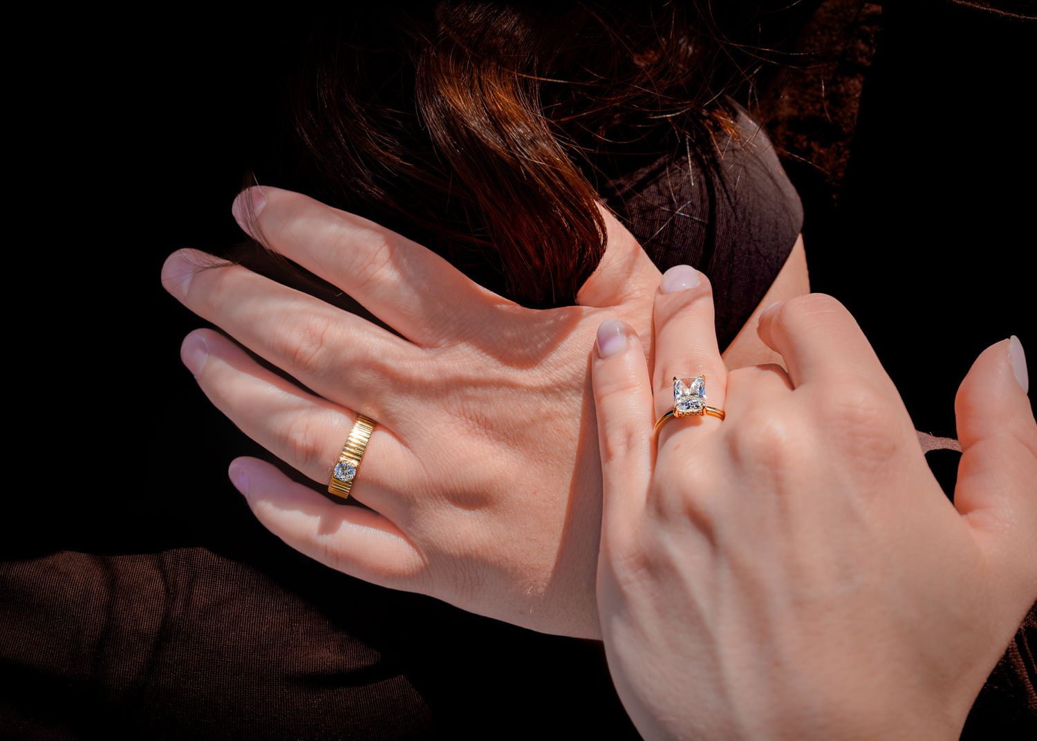 Two hands displaying wedding and engagement rings, one gold band and one diamond solitaire, against a dark background.