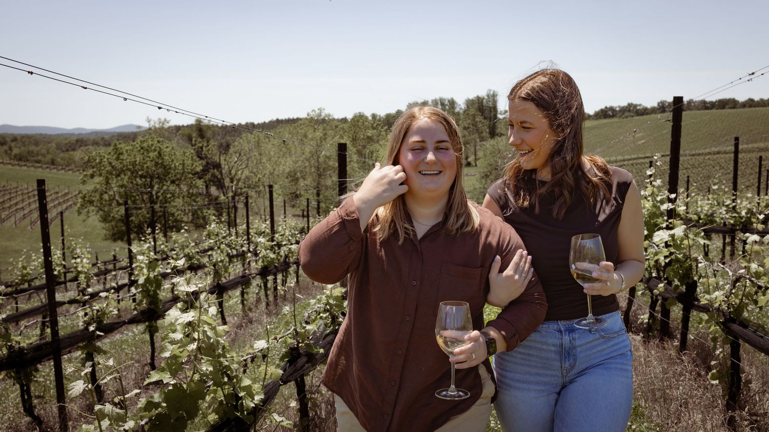 Two women enjoying wine glasses while walking through lush vineyard rows on a sunny day.