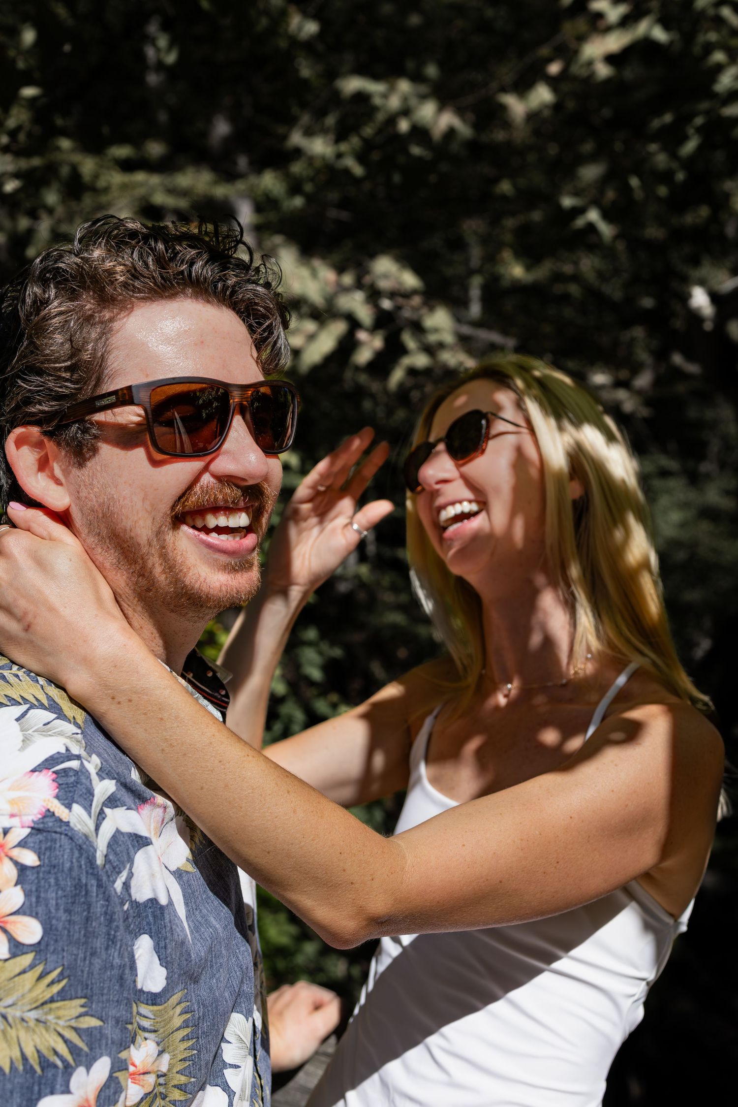 A joyful couple in sunglasses share a laugh together outdoors on a sunny day.