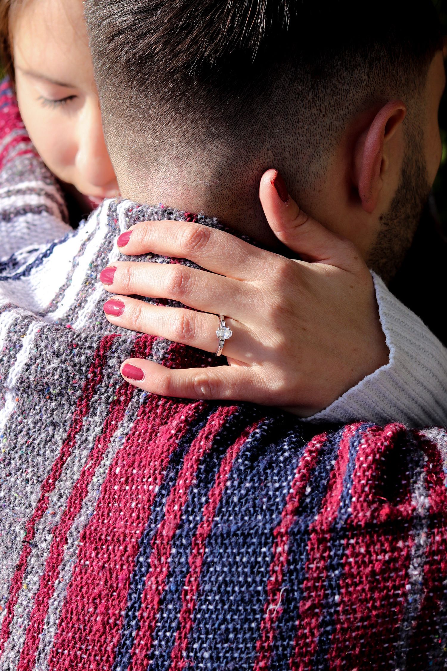 Close-up of a woman's hand with red nails and diamond engagement ring resting on a man's shoulder wrapped in a plaid blanket.