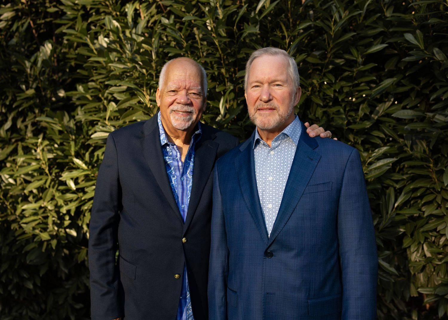 Two men in suits share a joyful moment while standing outdoors against a green foliage backdrop.