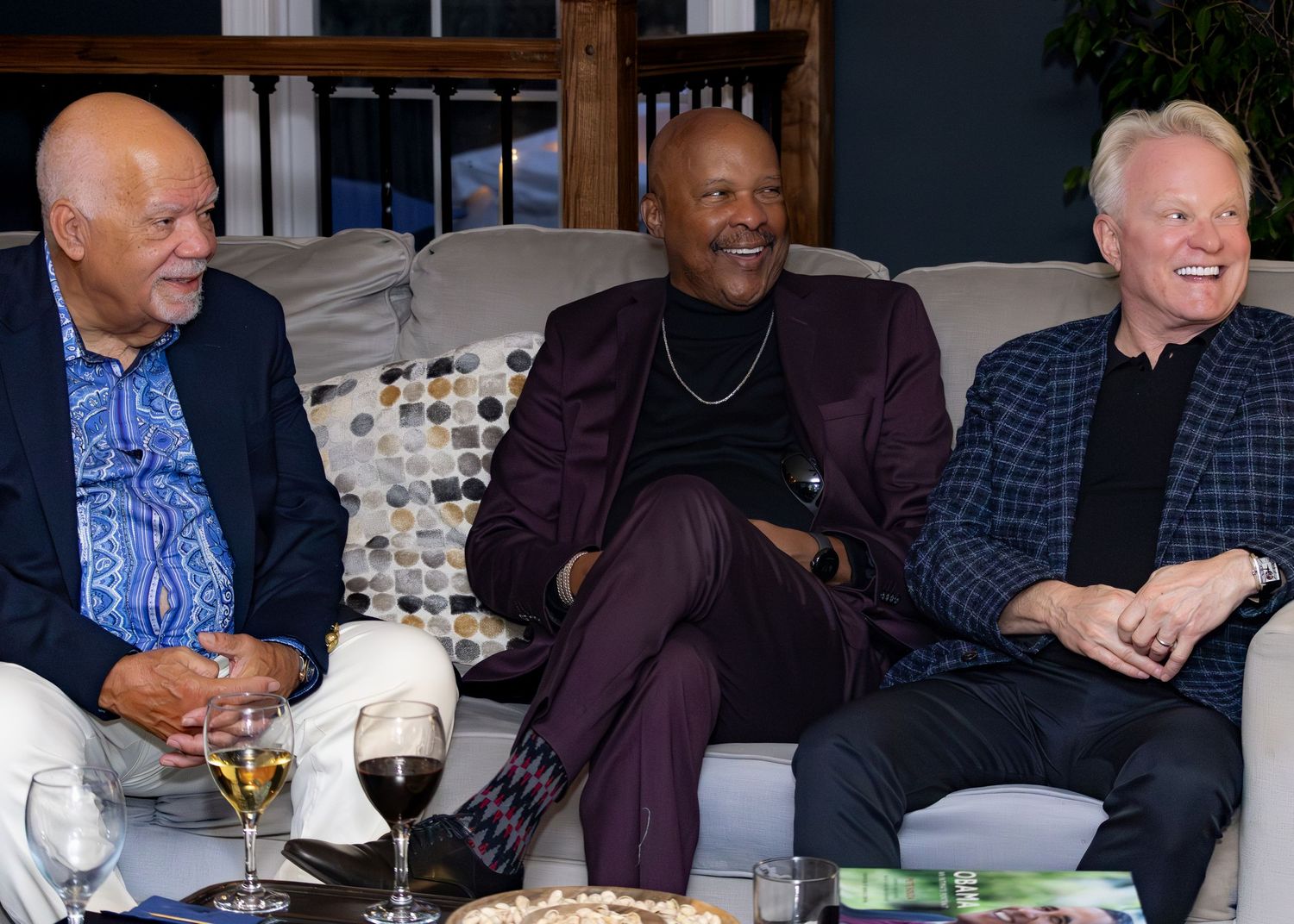 Three men in casual attire share laughs and drinks while sitting together on a couch at a social gathering.