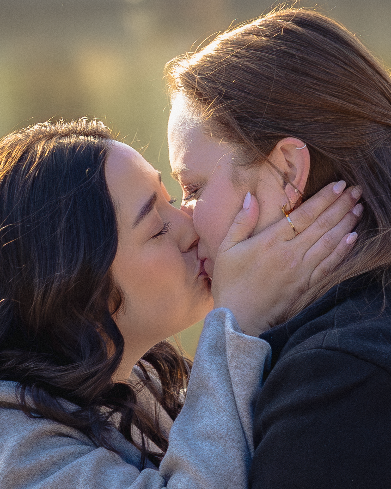 Two women sharing a romantic kiss outdoors in warm golden light, one with dark hair, one with auburn hair.