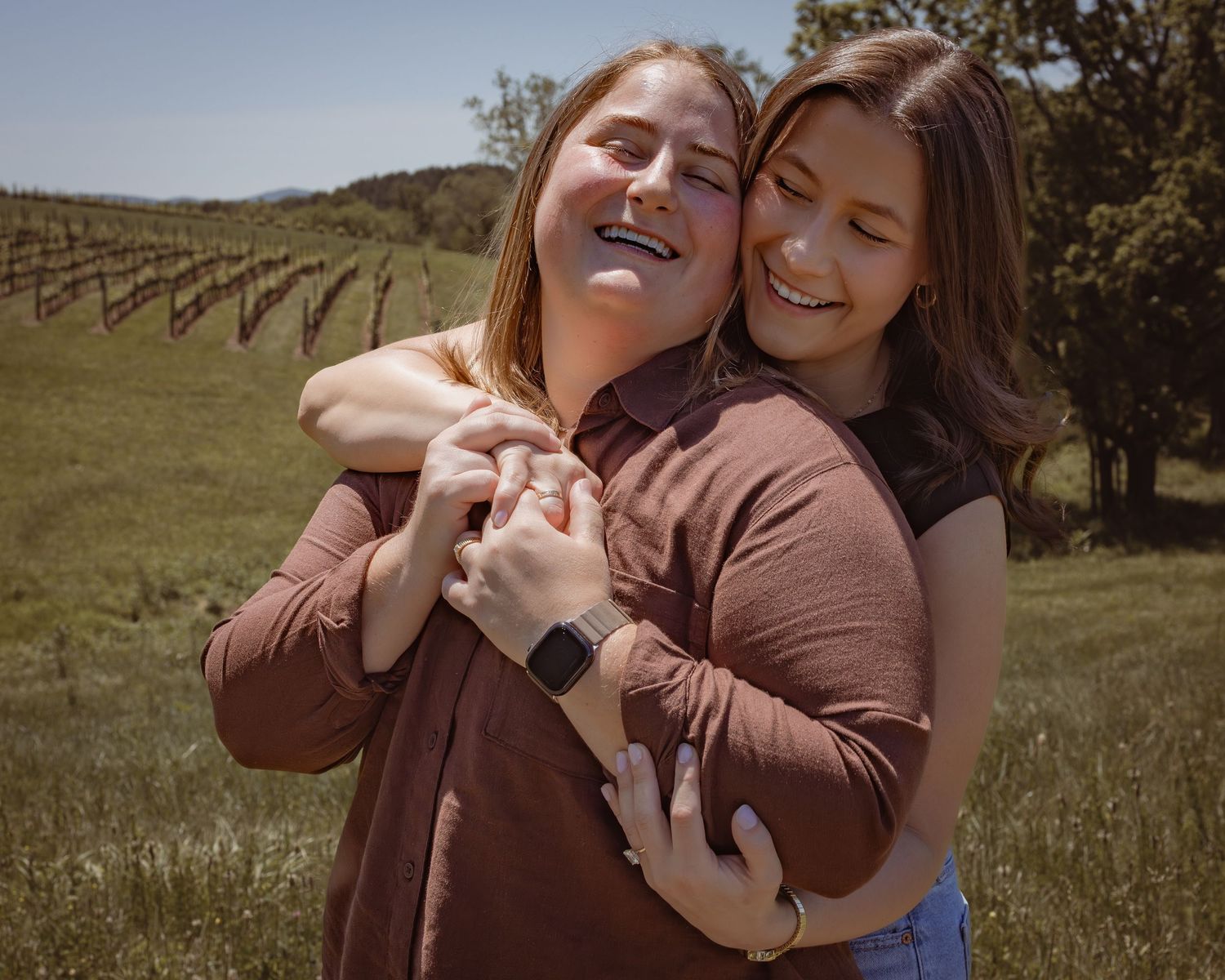 Two women hugging and smiling in a vineyard field, wearing brown tops on a sunny day.