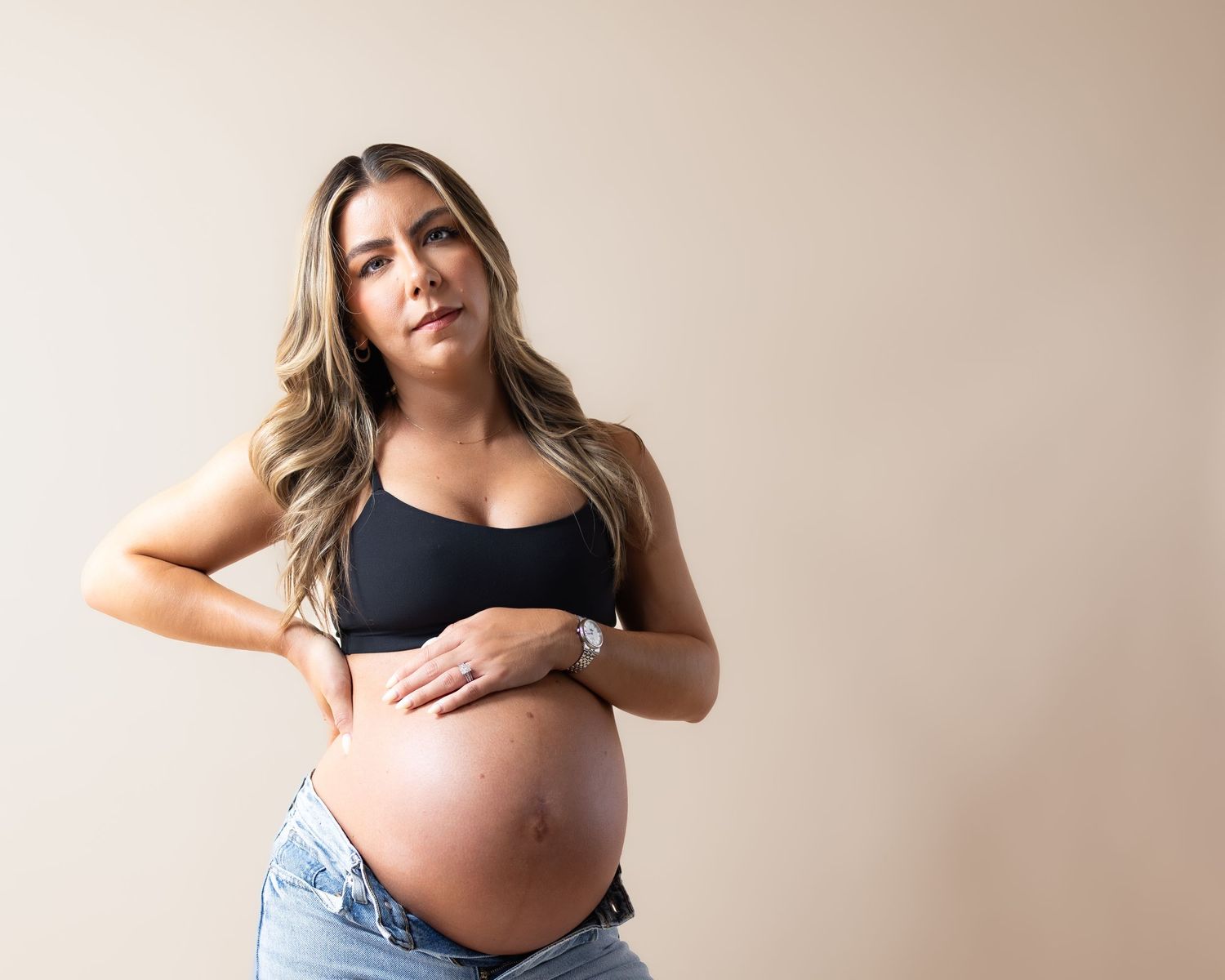 Pregnant woman in black crop top and jeans cradling baby bump against beige background.