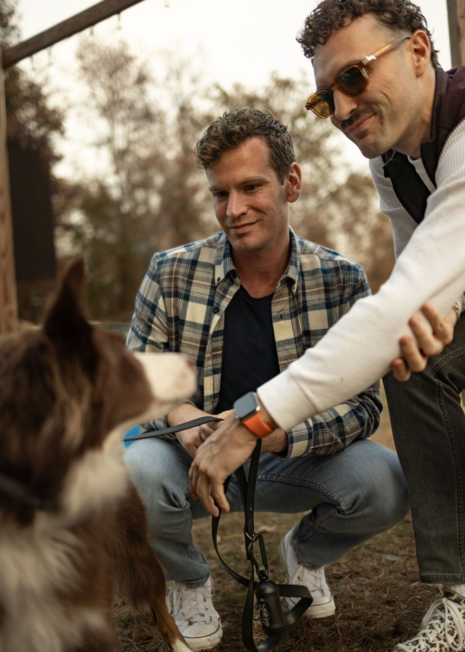 A person in a plaid shirt and sunglasses feeds a dog treats while sitting outdoors.