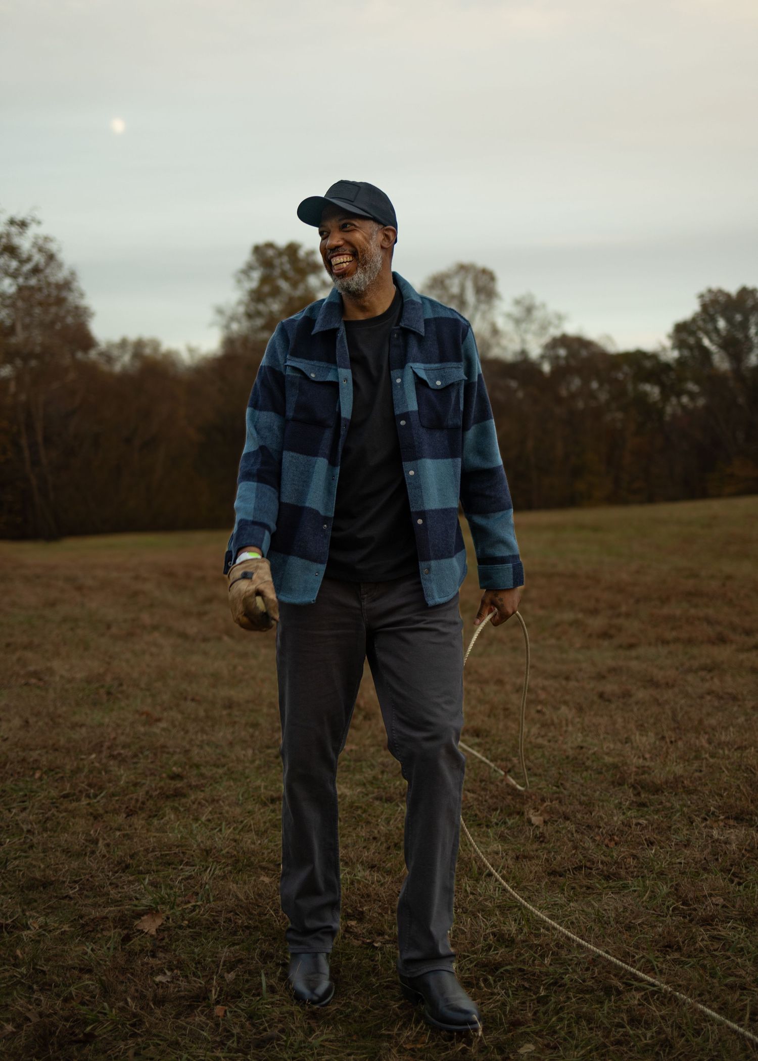 Person wearing a blue plaid flannel jacket and baseball cap stands in an autumn field at dusk.