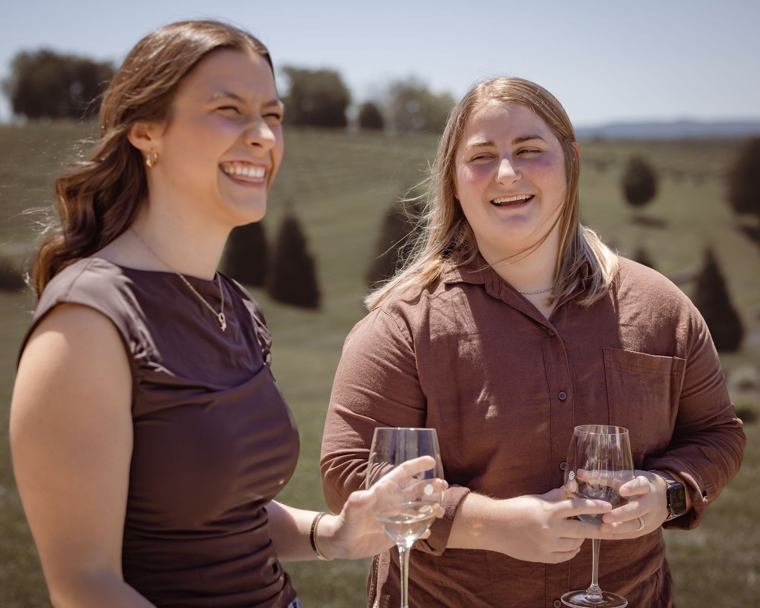 Two women smiling and holding wine glasses outdoors at a vineyard on a sunny day.