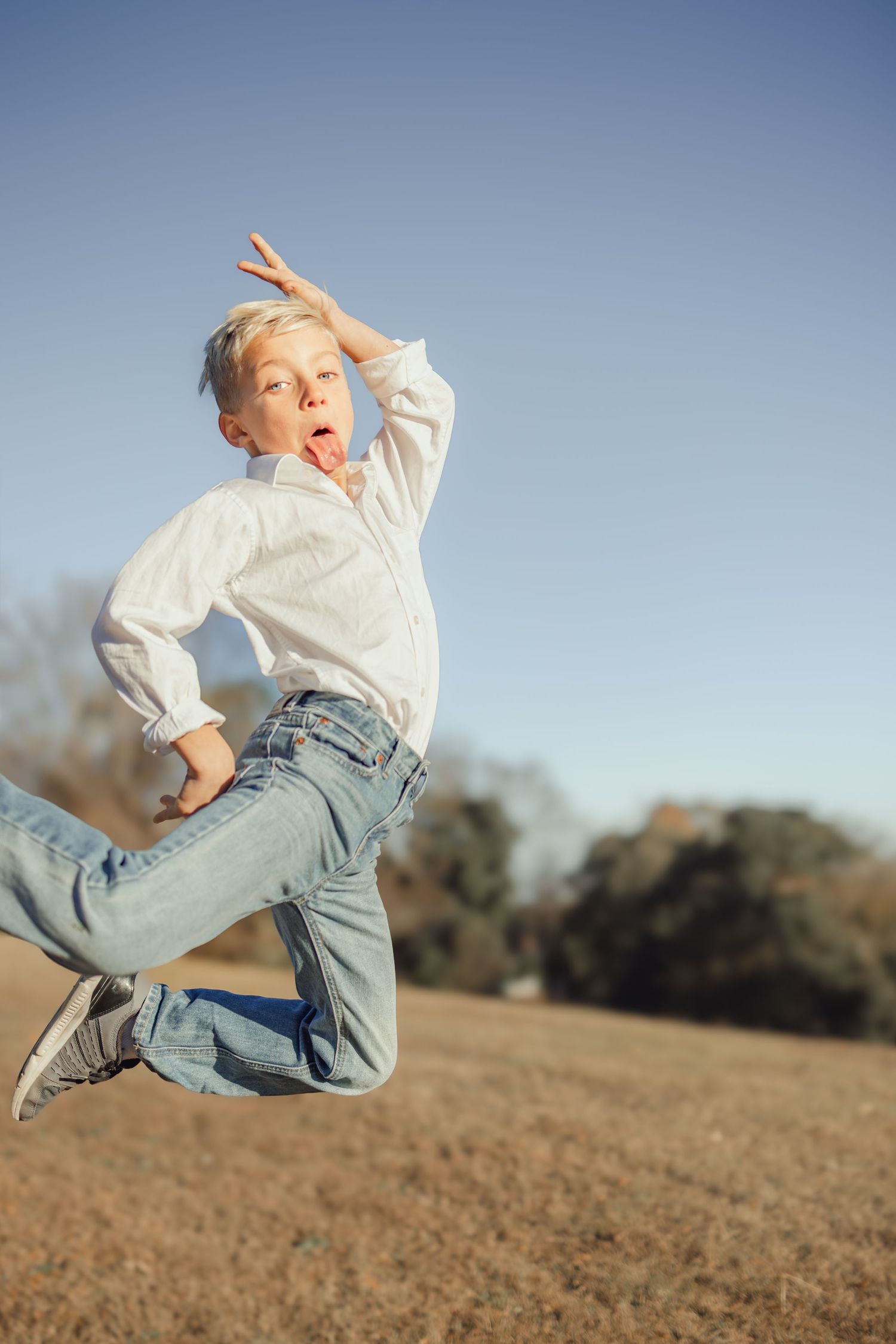 A young boy in a white shirt and jeans jumps energetically outdoors under a clear blue sky.