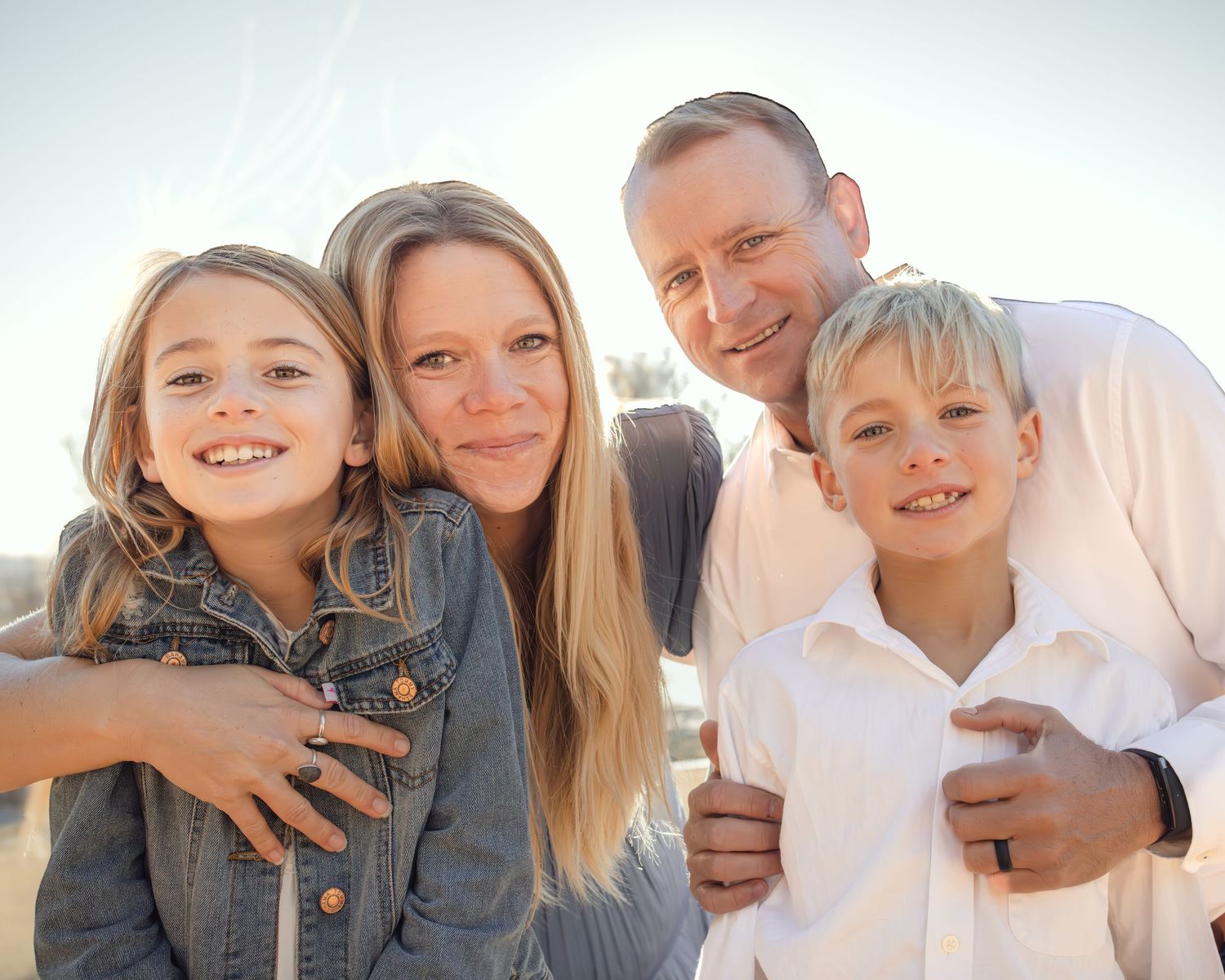 A happy family of four poses together outdoors in warm sunlight with bright smiles.