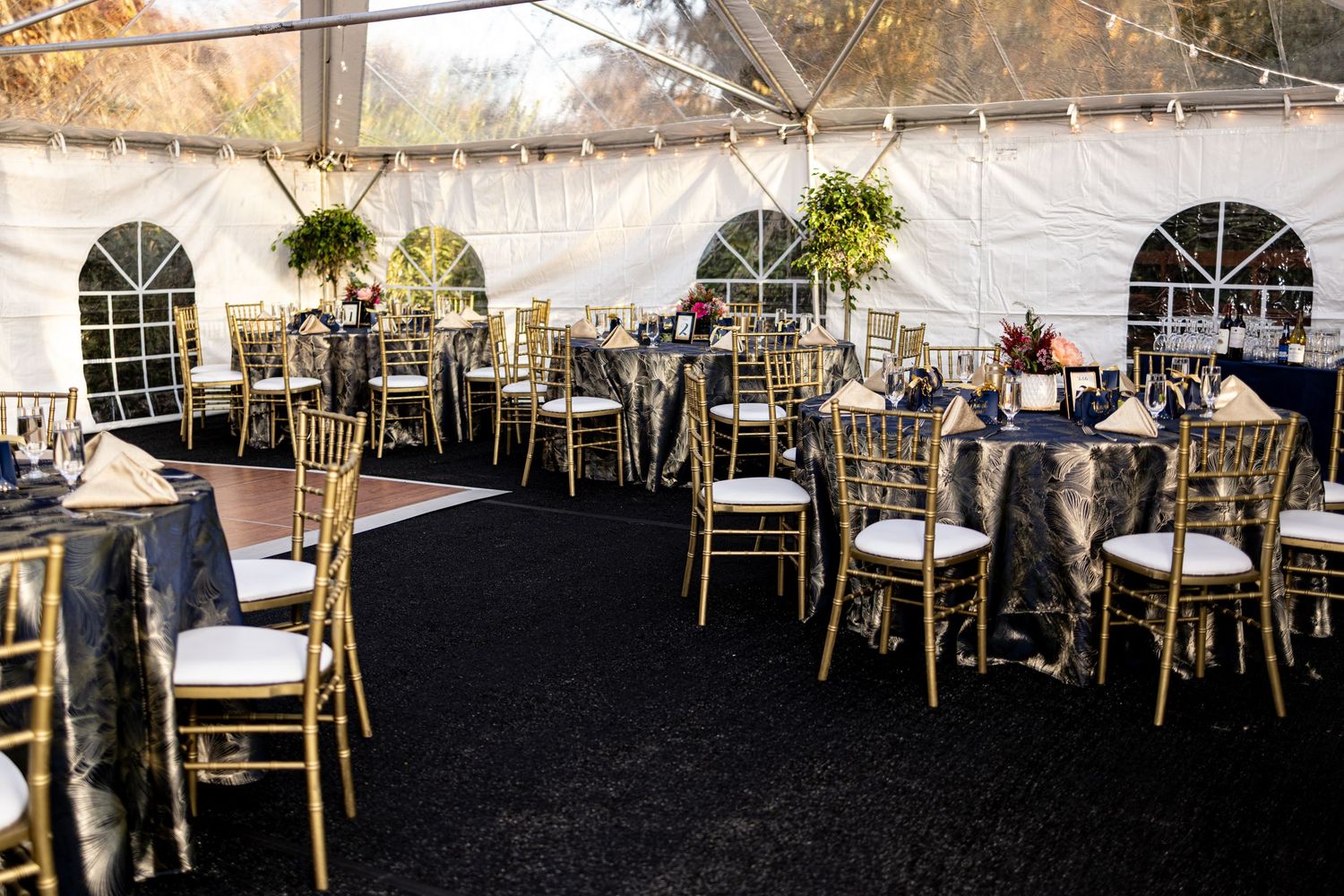 Wedding reception venue with elegant table settings under a white tent with chandelier lighting and gold chairs.