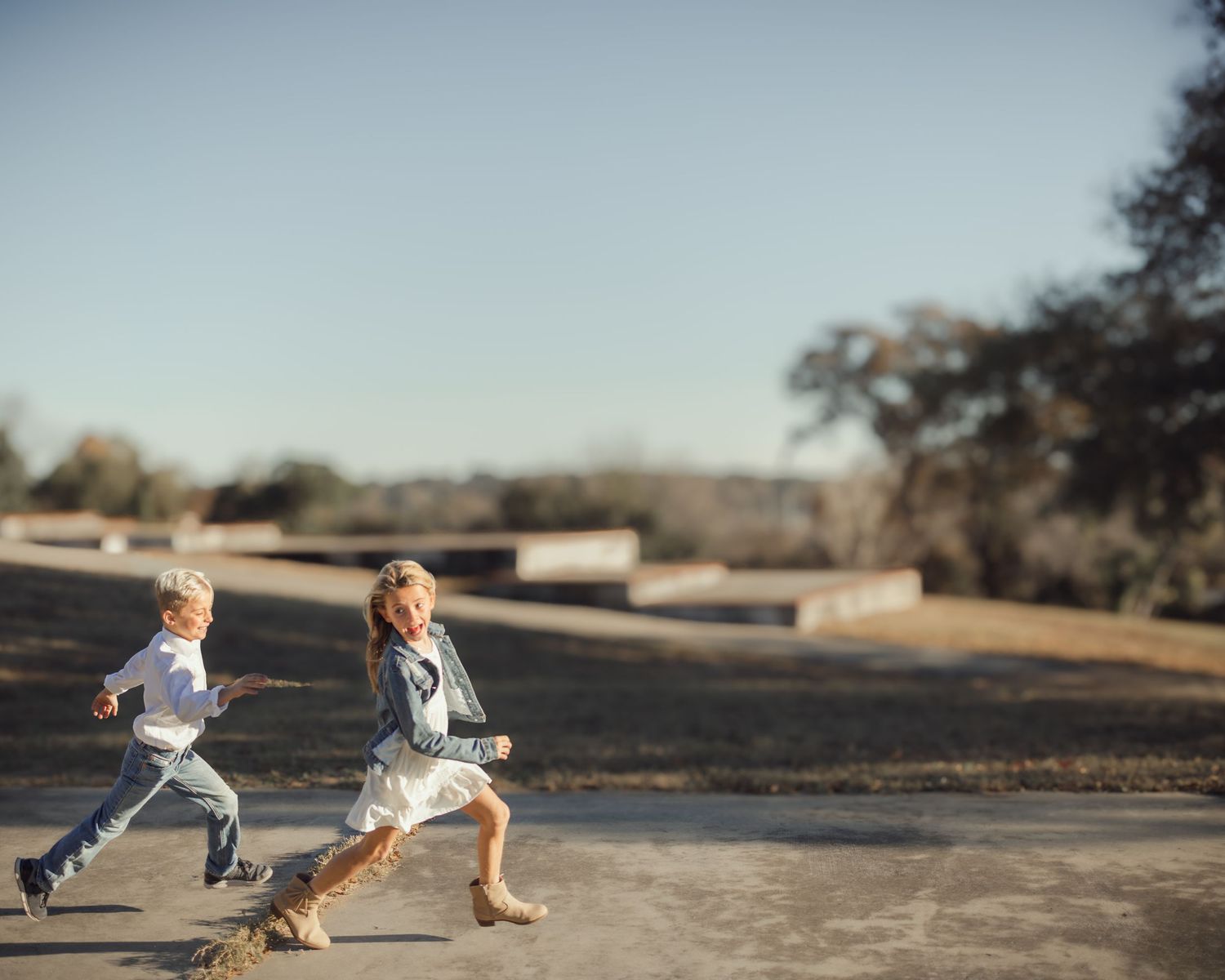 Two young children run together on a dirt path during a sunny afternoon in an outdoor setting.