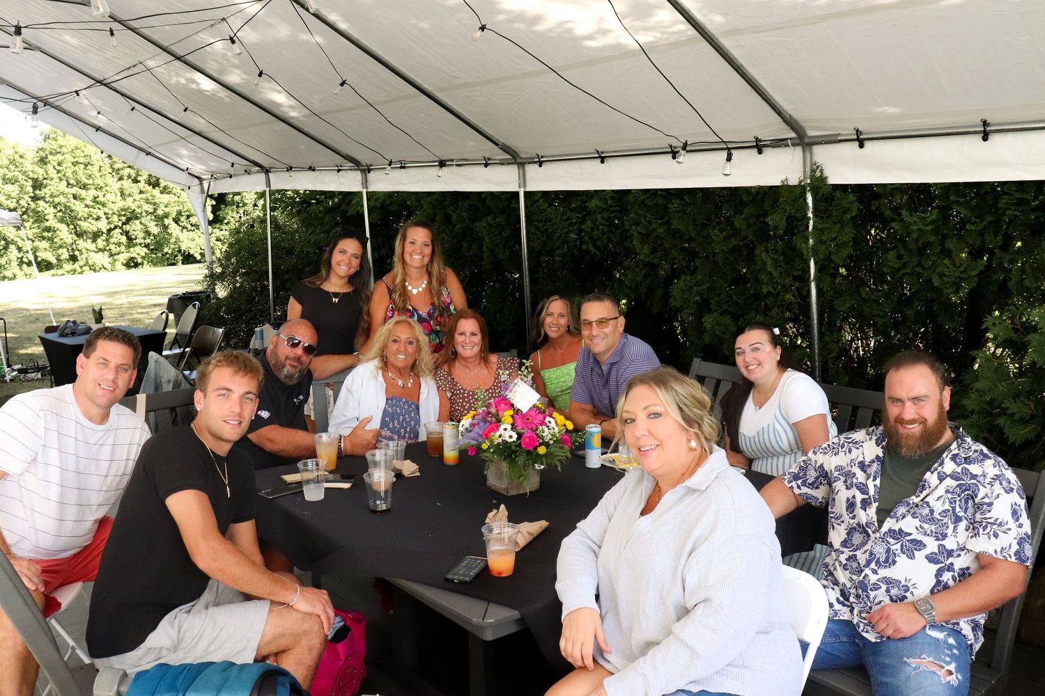 People gathered around outdoor tables at a social event enjoying food and drinks in the sunshine.