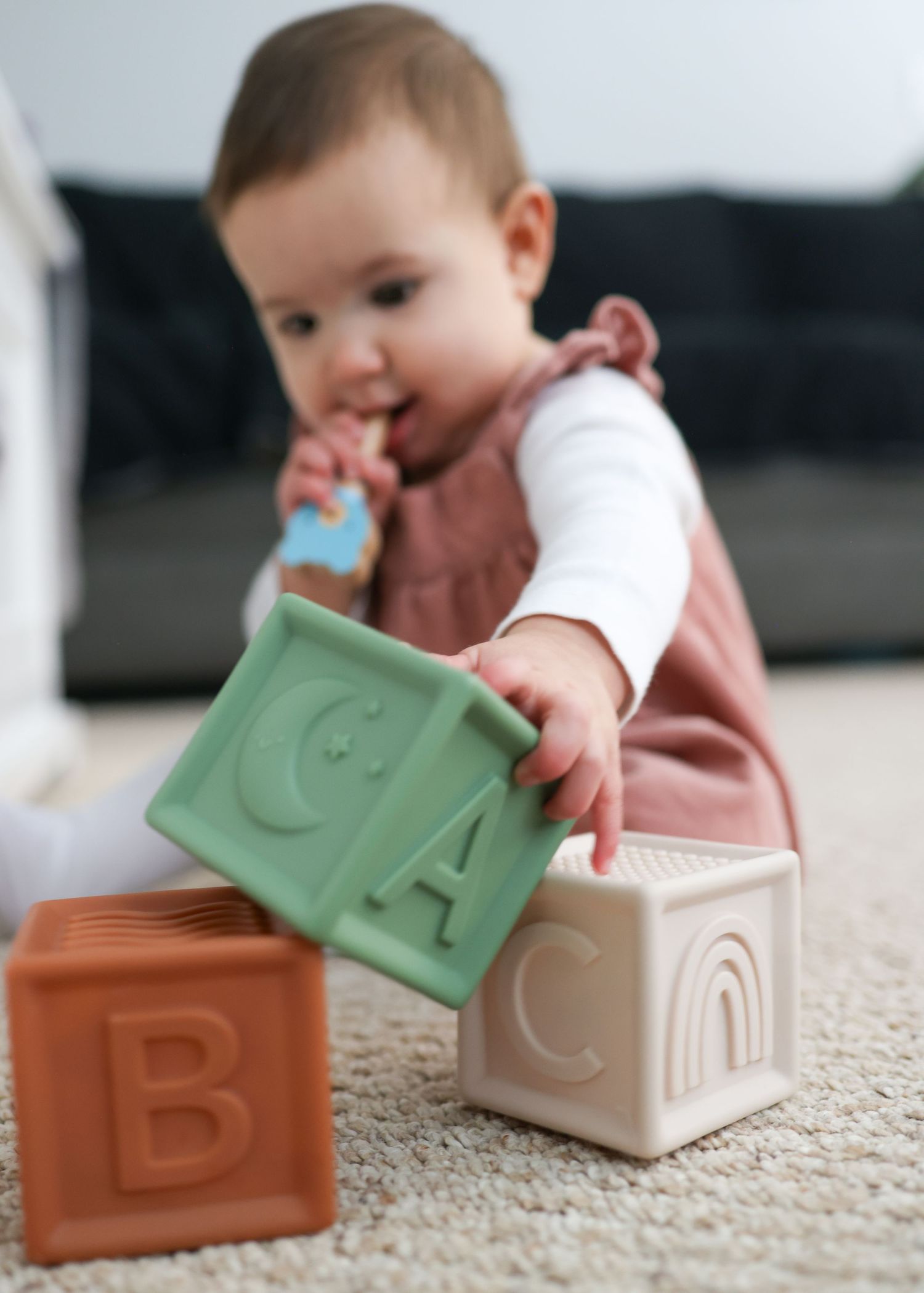 A baby plays with colorful alphabet building blocks arranged in a row on a beige carpet.