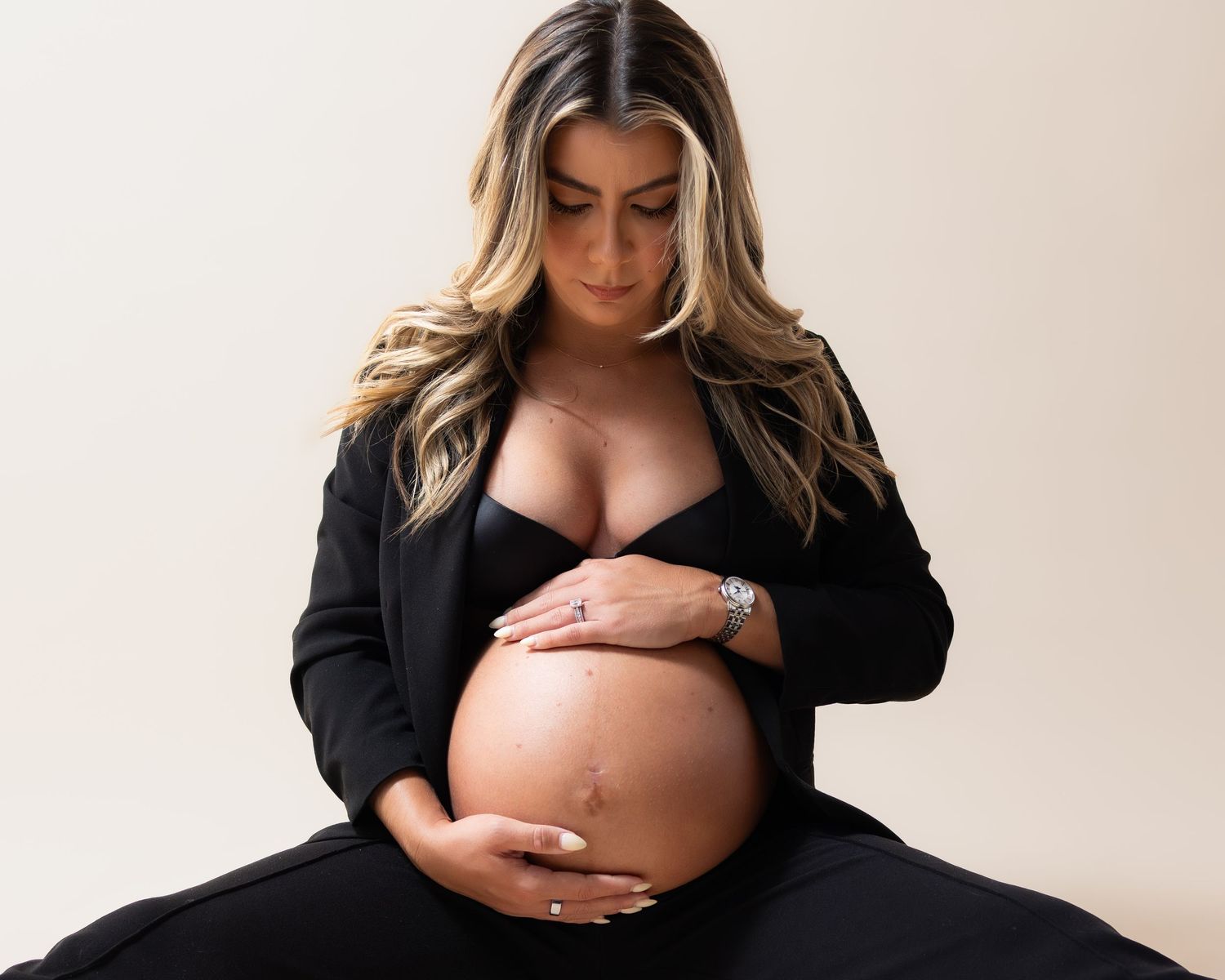 Pregnant woman in black outfit cradling her baby bump during a maternity photoshoot against a neutral background.