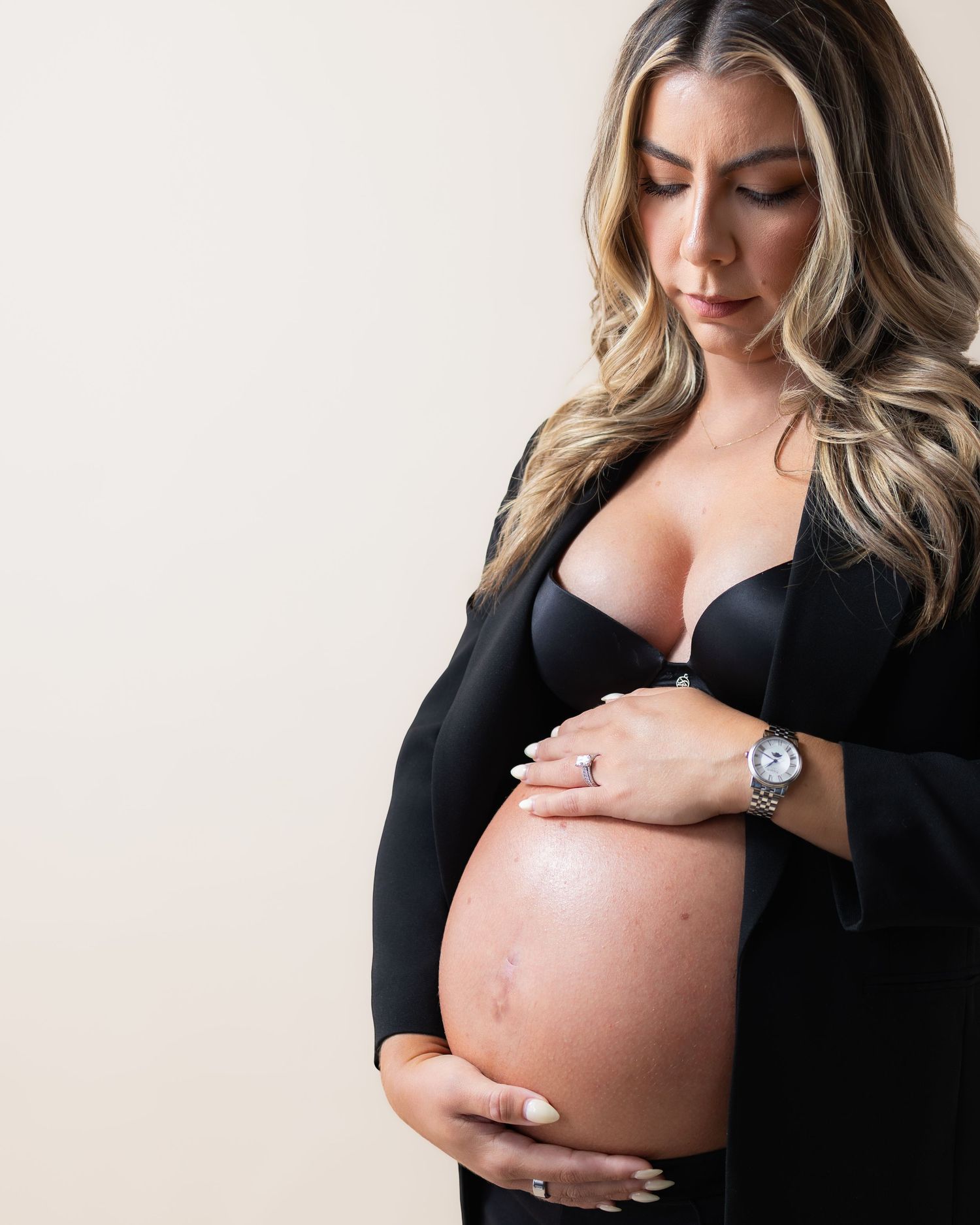 Pregnant woman in black outfit cradling her bare baby bump, wearing a silver watch and rings, with long blonde hair.