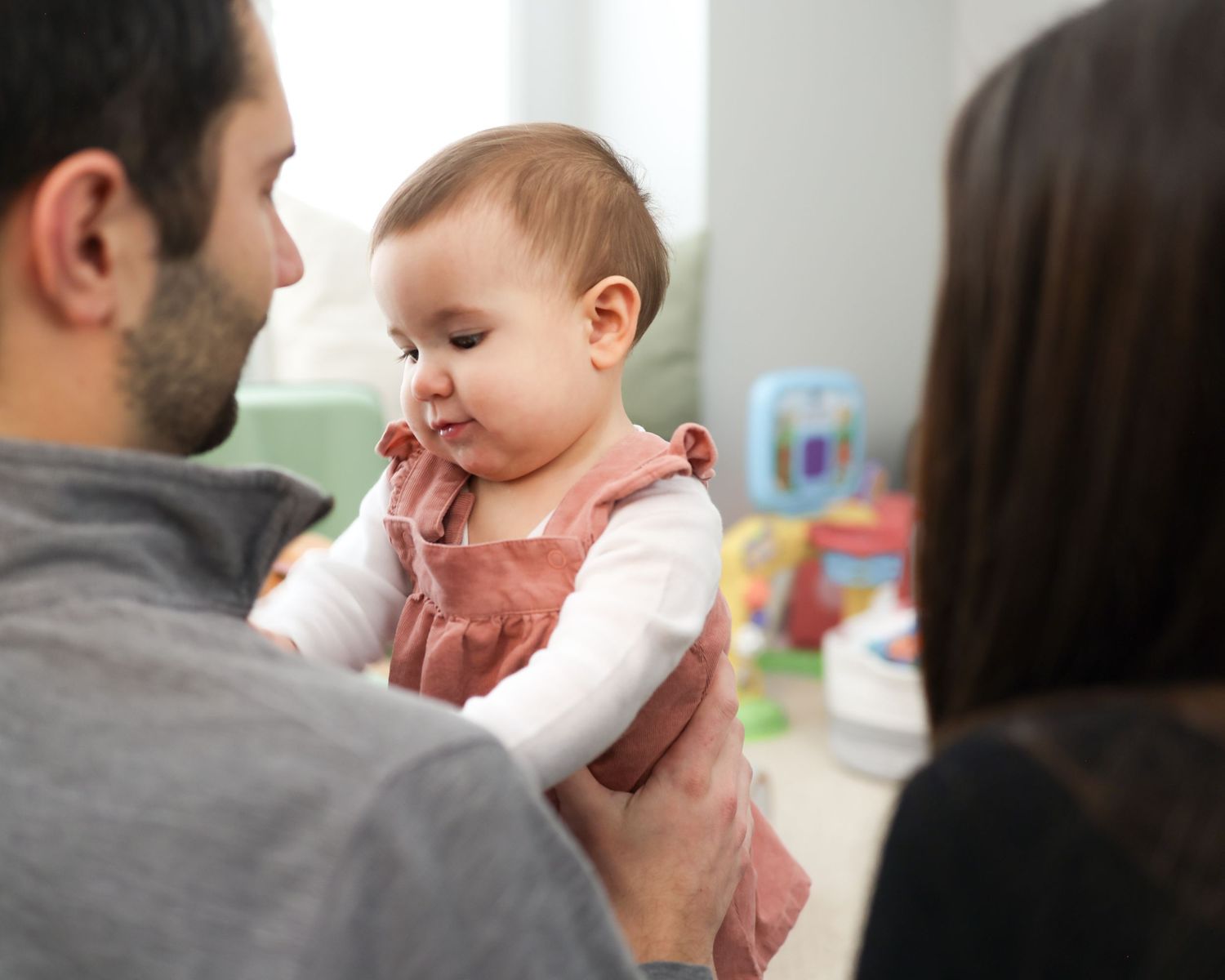 A father holds his adorable baby girl while a mother looks on in a bright, toy-filled nursery room.