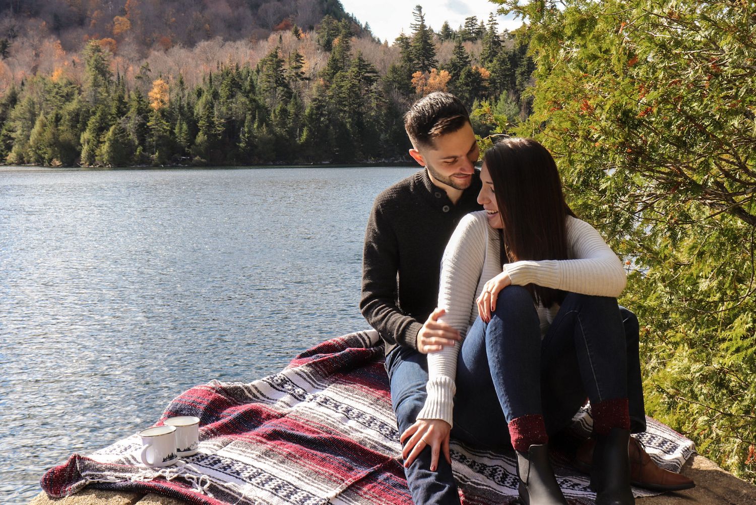 Couple sitting on a rocky lakeside ledge with a blanket, surrounded by autumn forest scenery.