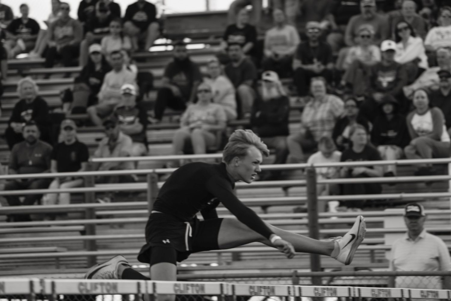Athlete clears a hurdle during a track and field competition in a packed stadium, captured in black and white.