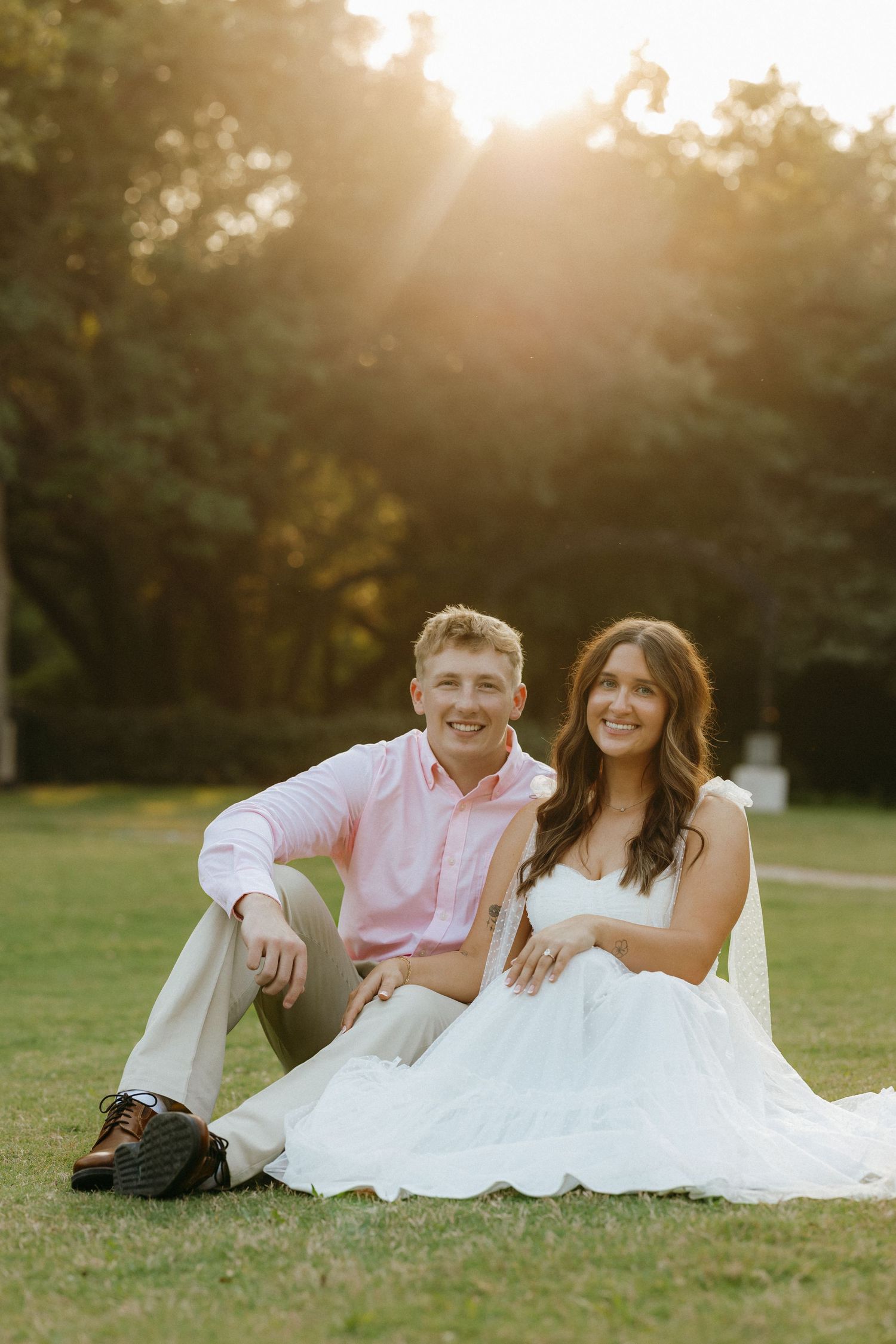 A smiling couple sits on green grass at golden hour, man in pink shirt and woman in white dress, with sunlit trees behind.
