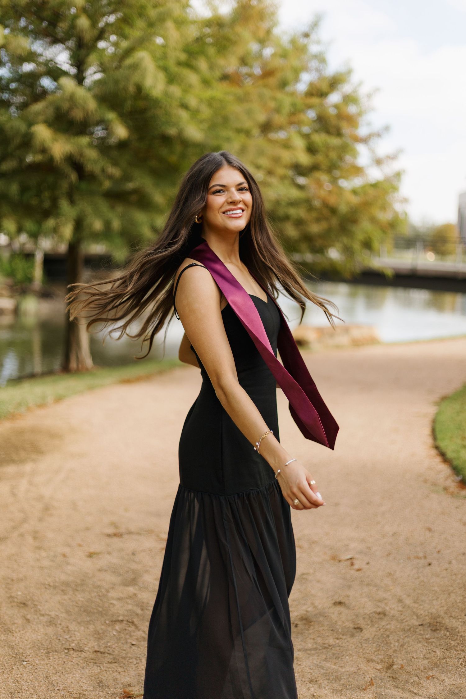 Woman in black dress twirling and dancing on a dirt path with trees in background.