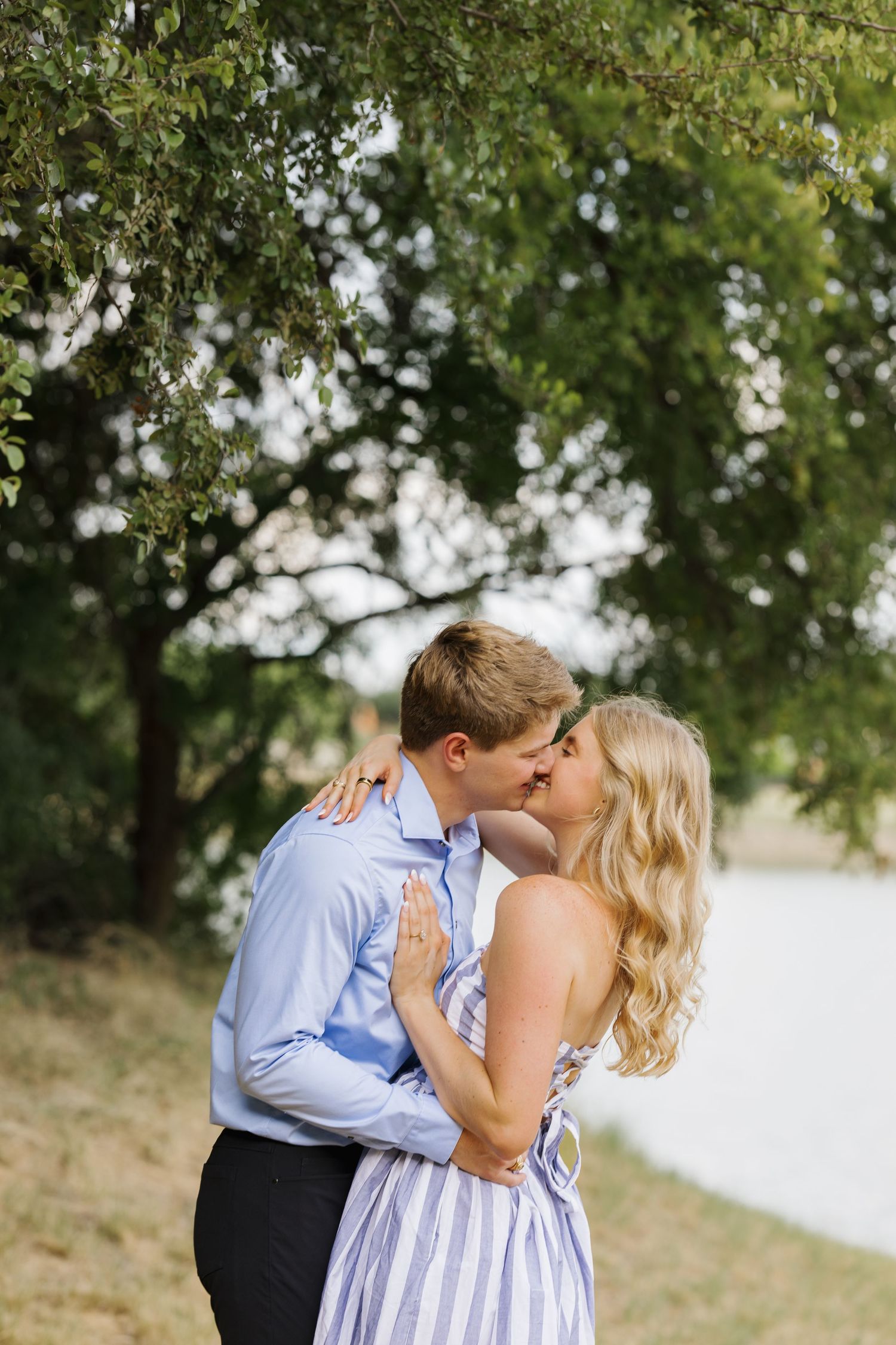 A couple shares a romantic kiss outdoors under a lush green tree, woman in striped dress, man in blue shirt.