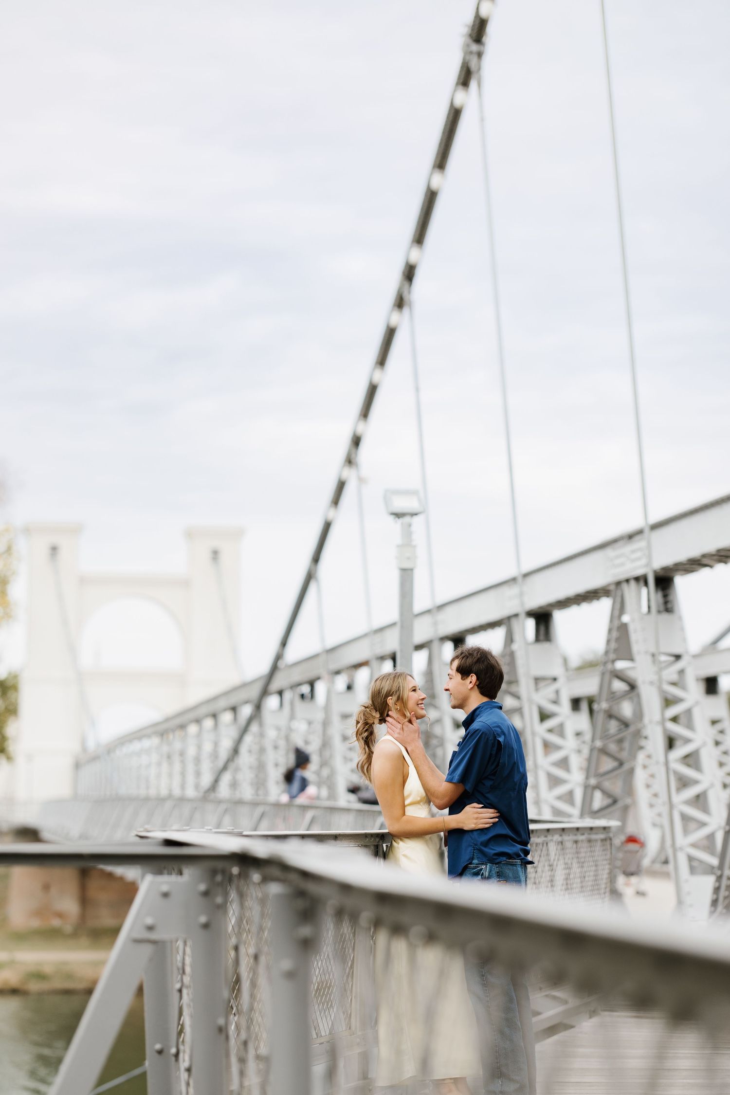 A couple embraces on a white bridge with architectural details visible in the background.