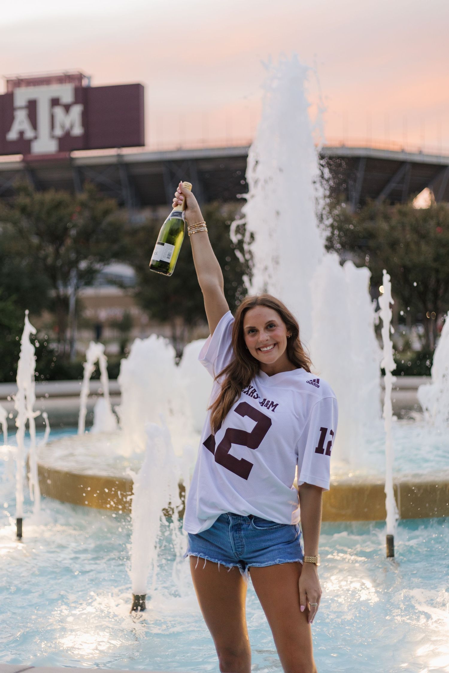 Graduate celebrates near fountain wearing Texas A&M shirt and denim shorts.