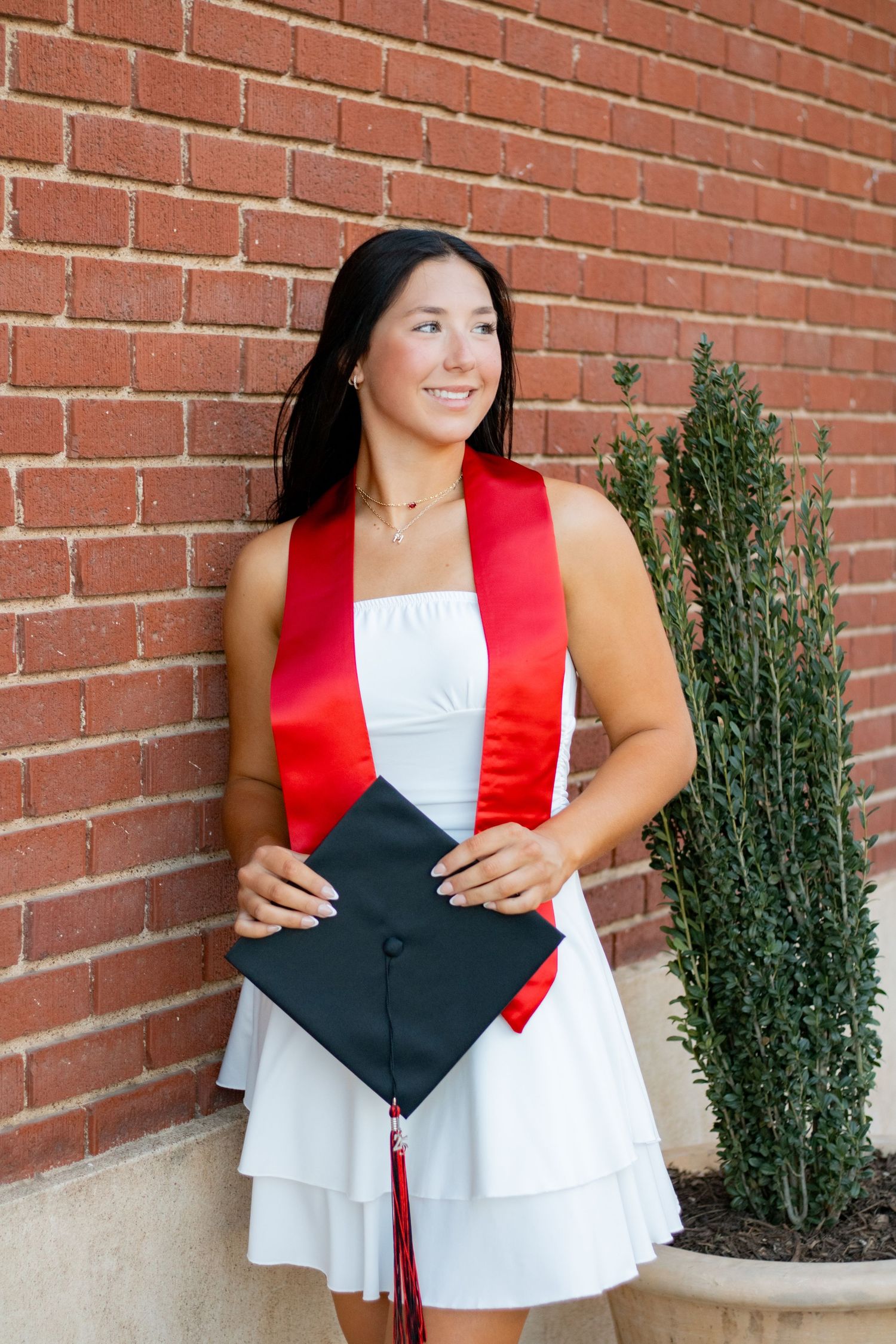 Graduate poses with cap against brick wall wearing white dress and red stole.