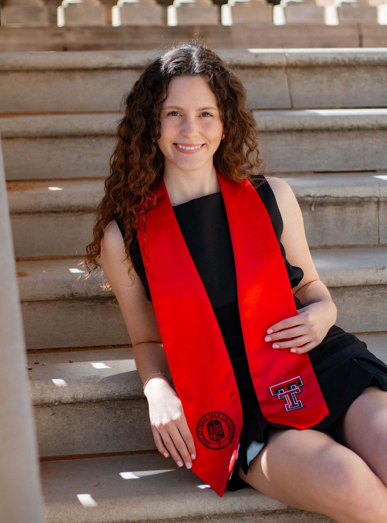 Student in black outfit and red graduation stole poses on stone steps in outdoor portrait session.