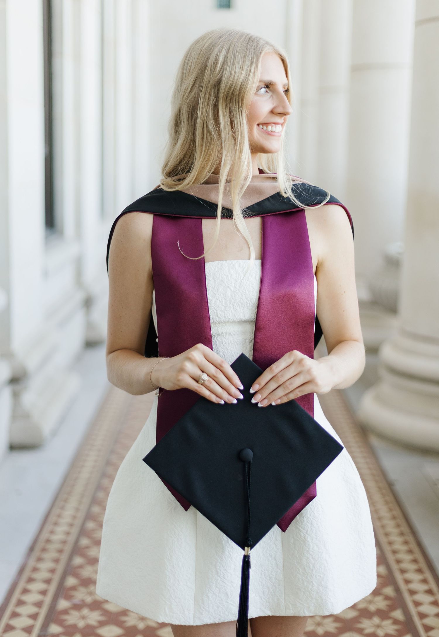 Graduate wearing white dress and burgundy stole posing in columned hallway.