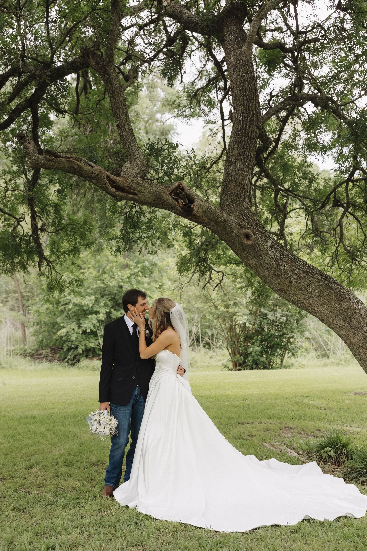 Bride and groom sharing a kiss under a large oak tree in a lush green garden on their wedding day.