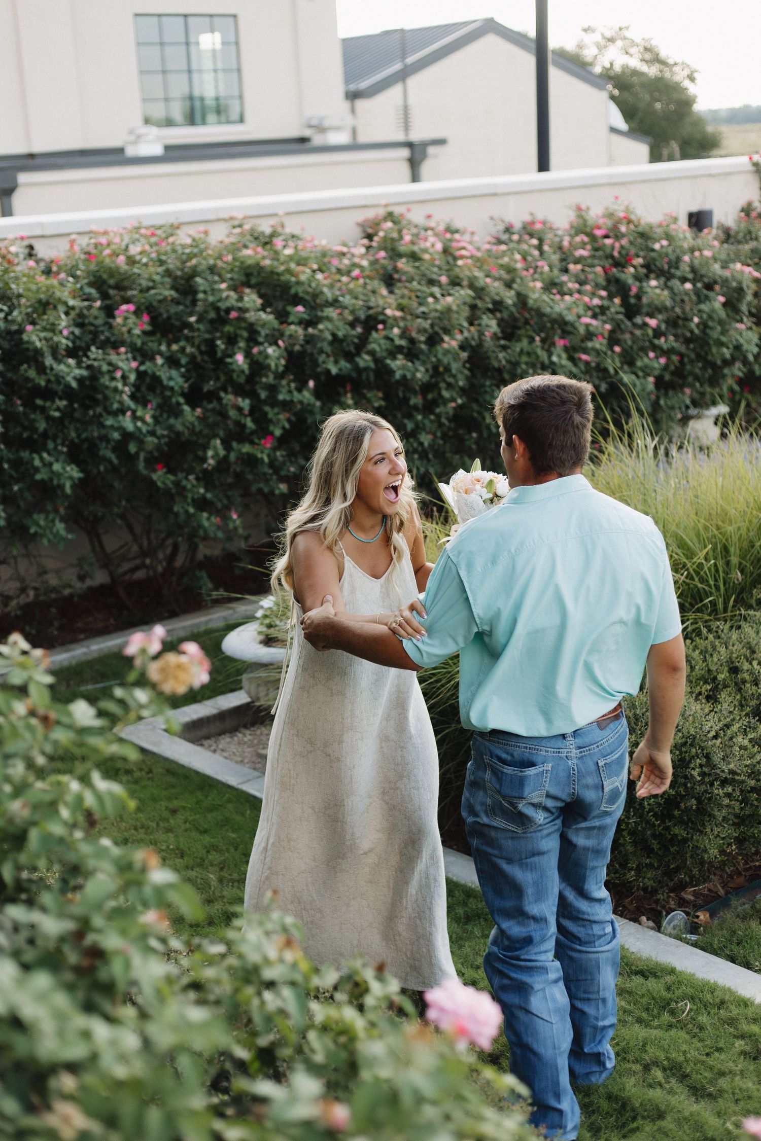 A couple walking through a garden with rose bushes and greenery during a casual outdoor photoshoot.
