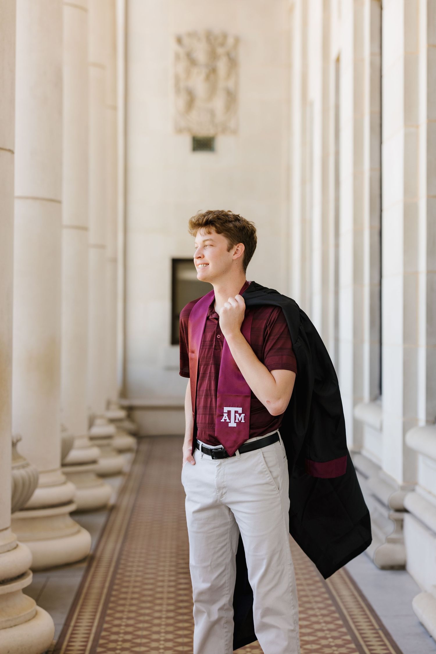 Student in maroon Texas A&M shirt and white pants carries a black jacket while walking through a classical columned hallway.