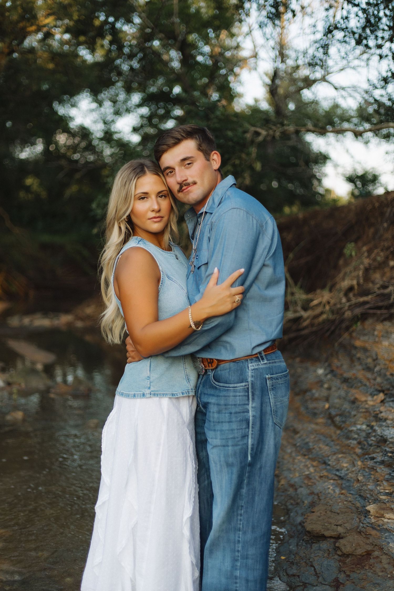 A couple sharing intimate moments by a rocky creek, with her in white dress and him in denim.