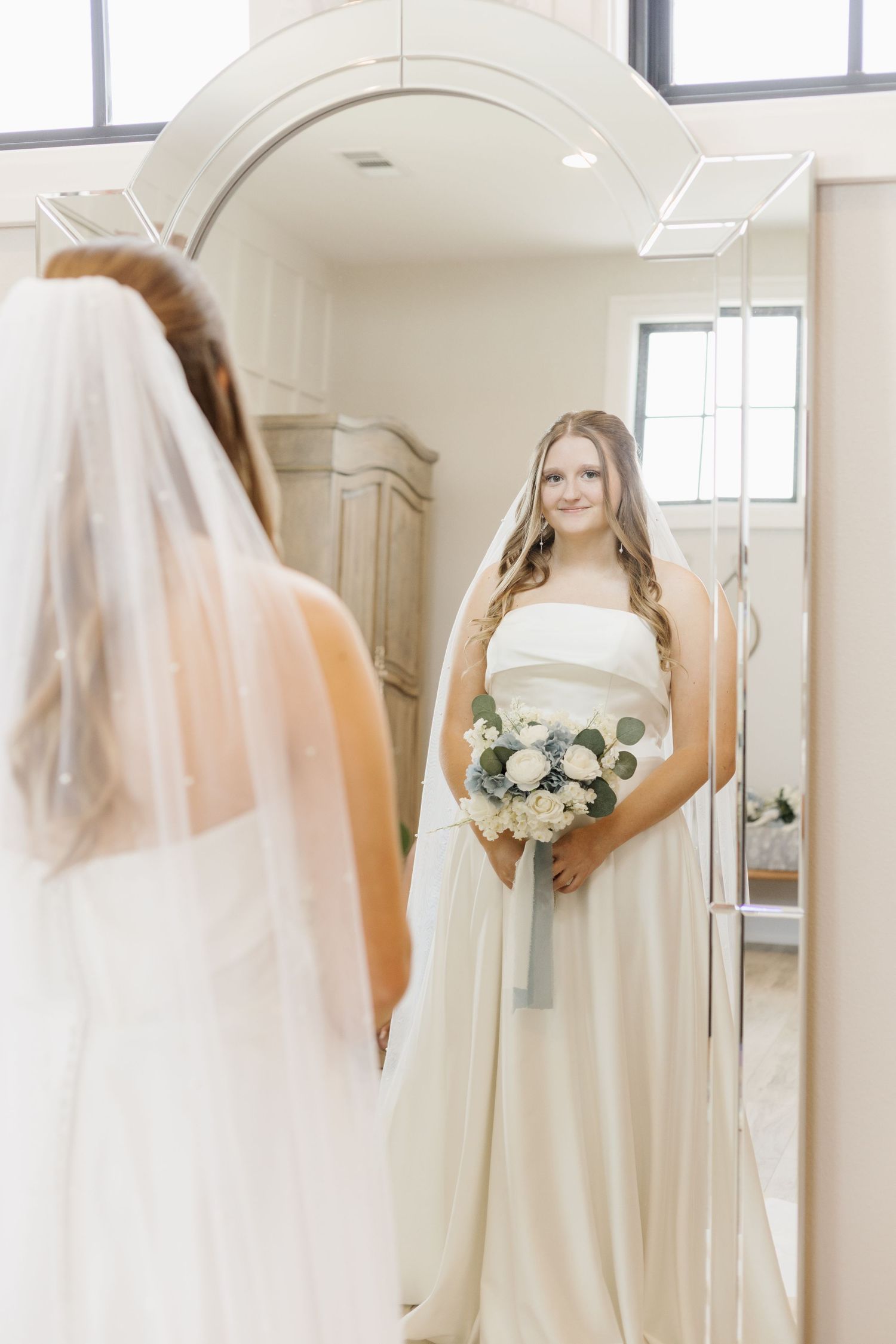 Multiple reflections of a bride in her wedding gown holding a white and green bouquet in a mirrored room.