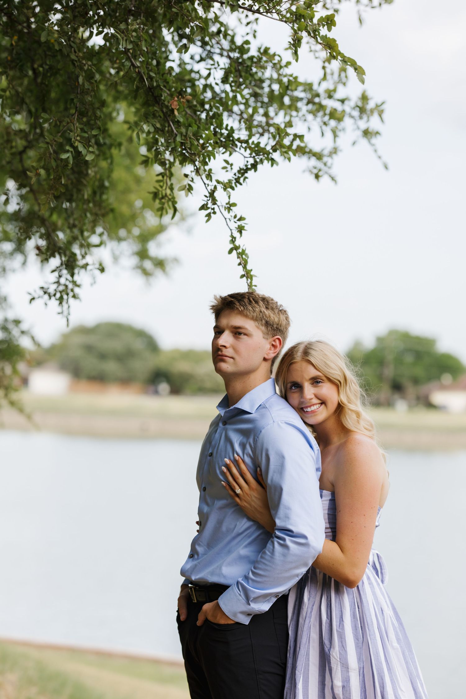 A young couple poses romantically by a lakeside under a tree, the woman in a lavender dress embracing the man in a blue shirt.