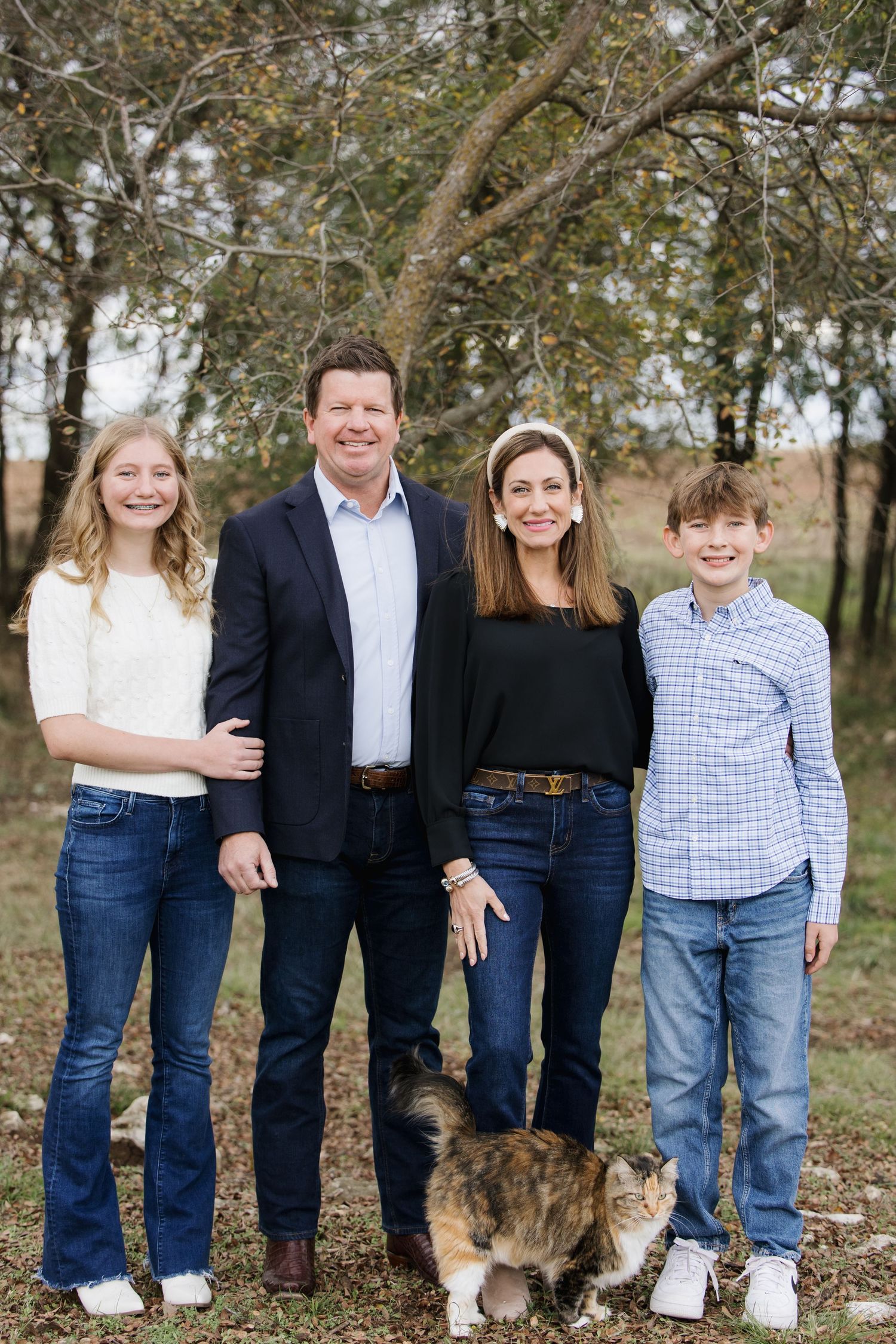Family portrait outdoors with a cat in autumn setting with trees in background.