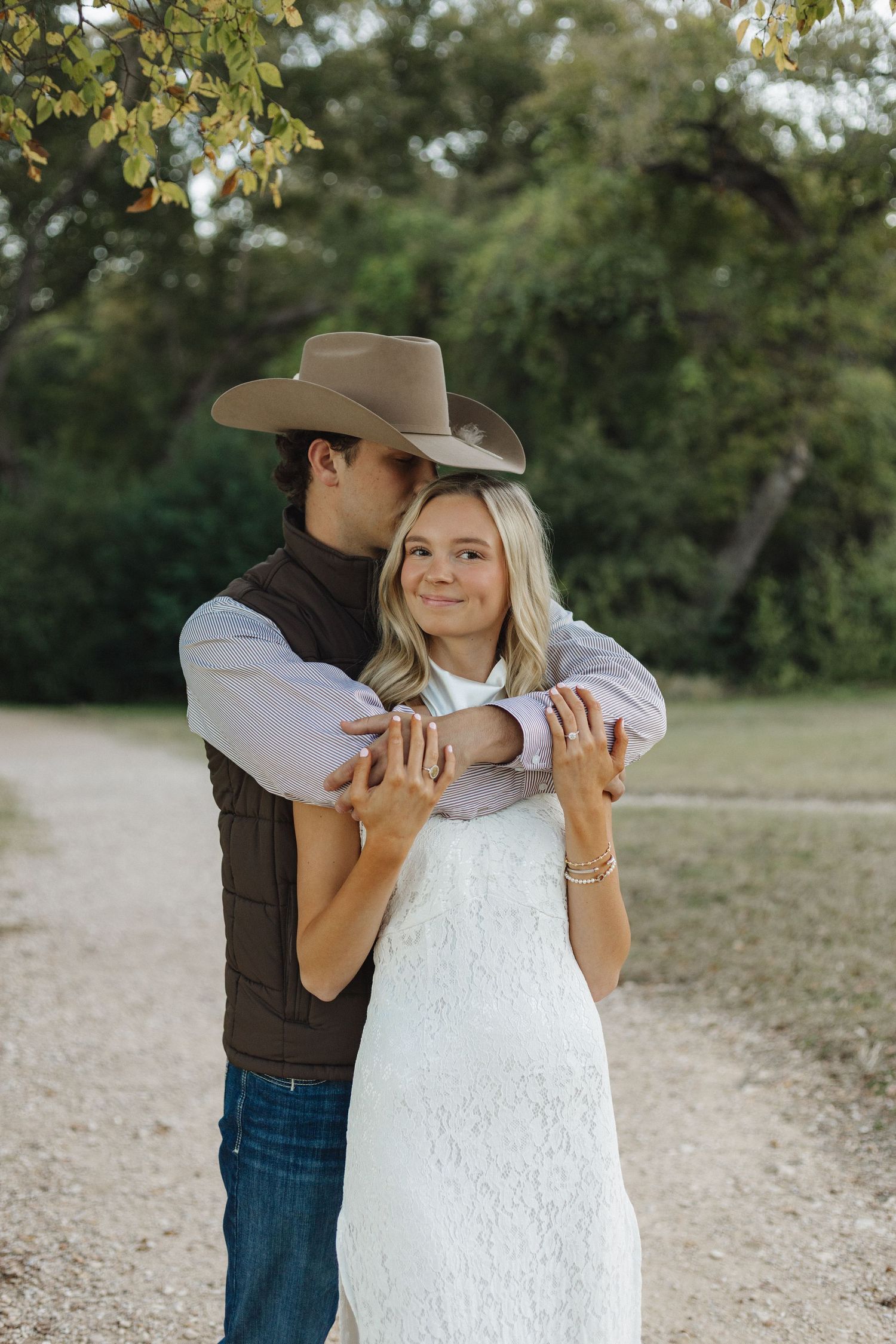Couple in western attire embracing on gravel path with trees in background.