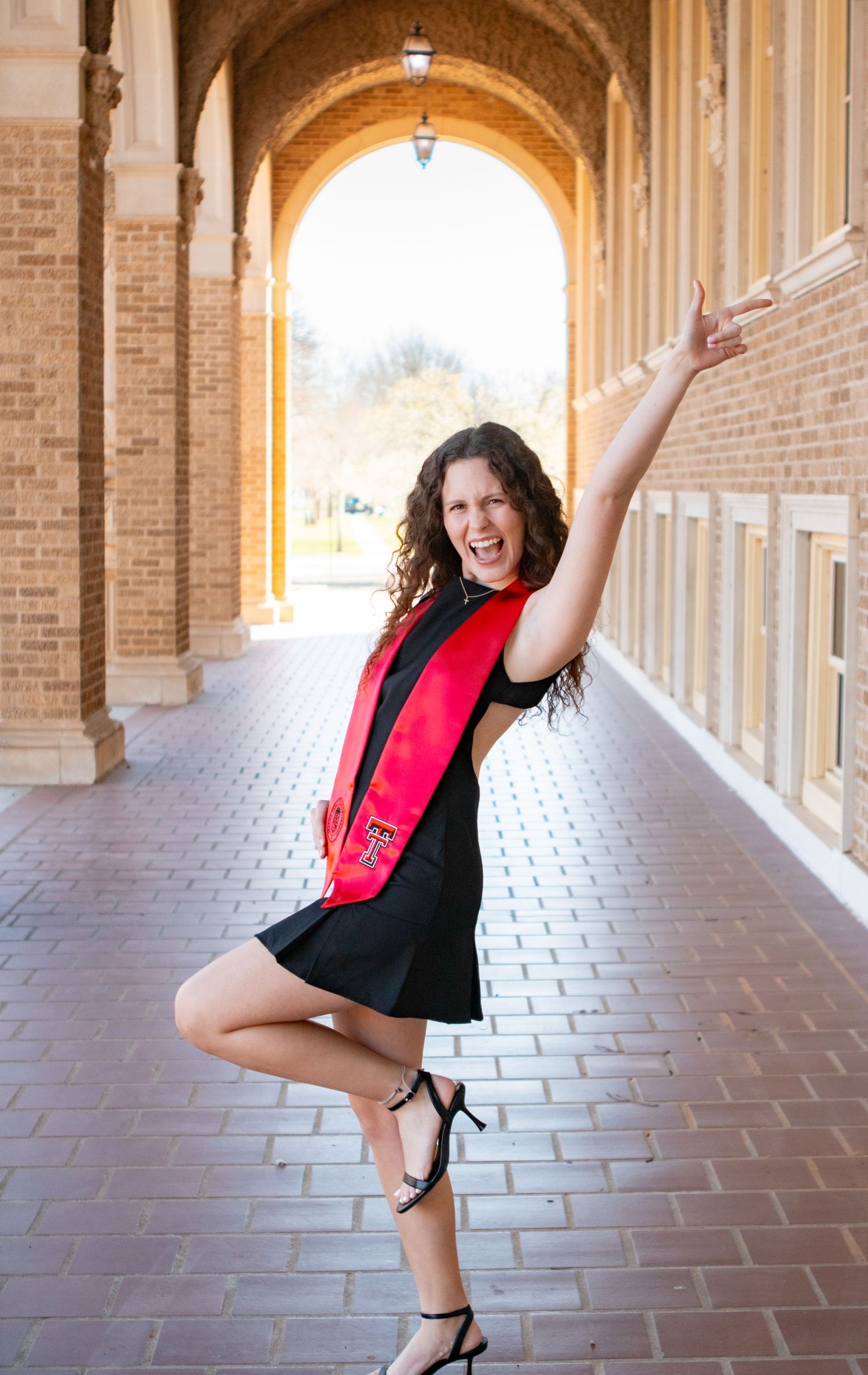 Graduate in black dress and red stole dances joyfully in arched university hallway.