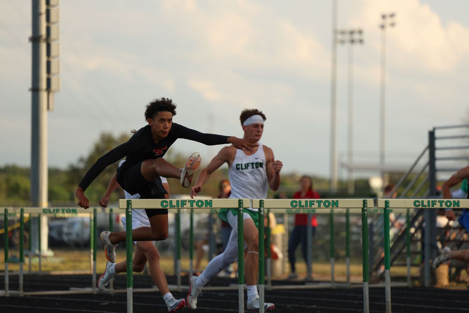 Two athletes compete in a hurdles race at a Clifton track and field event under cloudy skies.