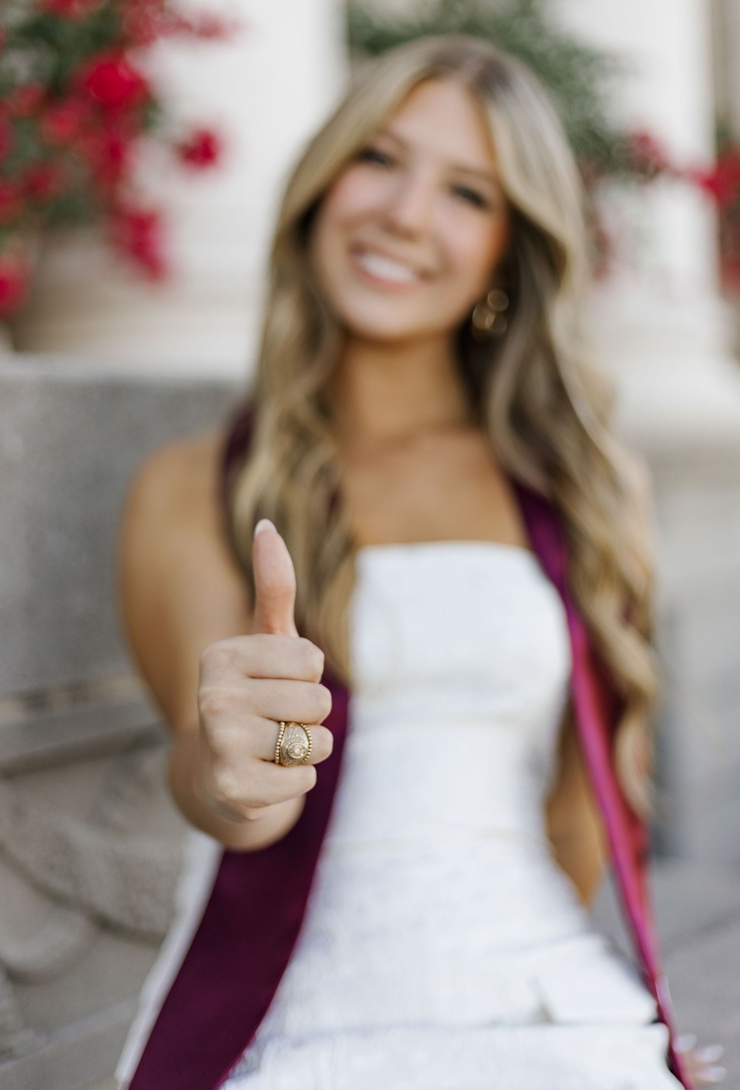 Graduate in white dress and burgundy stole giving thumbs up near red flowers.